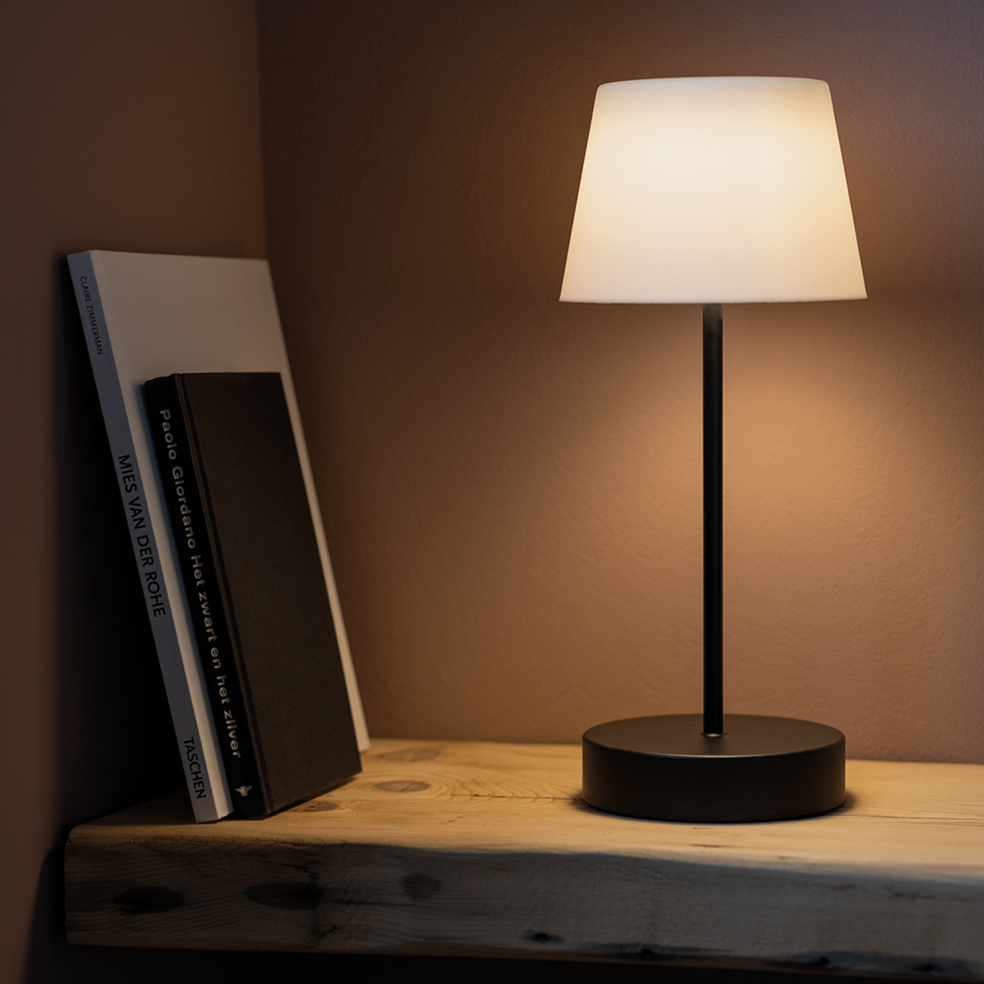 Eine 'pure' Oscar Tischleuchte steht auf einem Holzbrett neben zwei Büchern. Die Lampe hat einen schwarzen Fuß und einen weißen Lampenschirm, der warmes Licht ausstrahlt.
