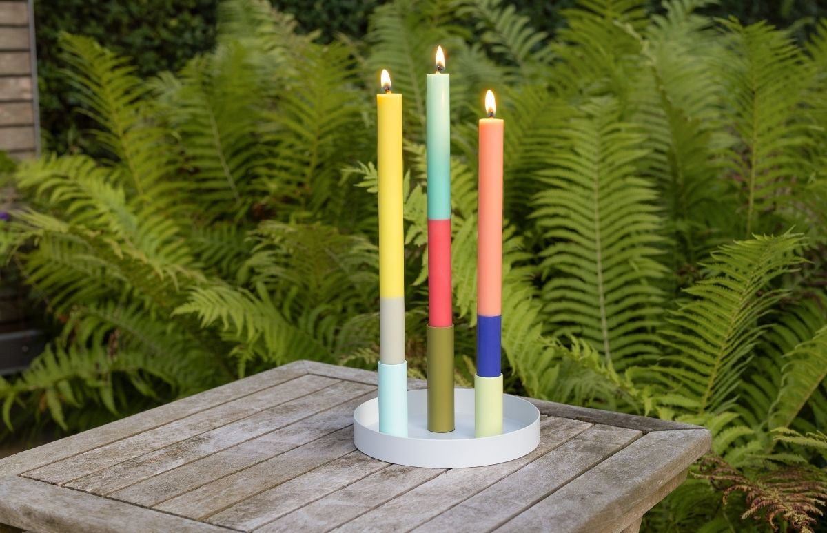 Trois bougies multicolores lumineuses dans un plat rond blanc sur une table de jardin en bois. À gauche : jaune, gris, bleu clair ; au centre : turquoise, rouge, vert olive ; à droite : orange, bleu foncé, vert clair. Fougères en fond.