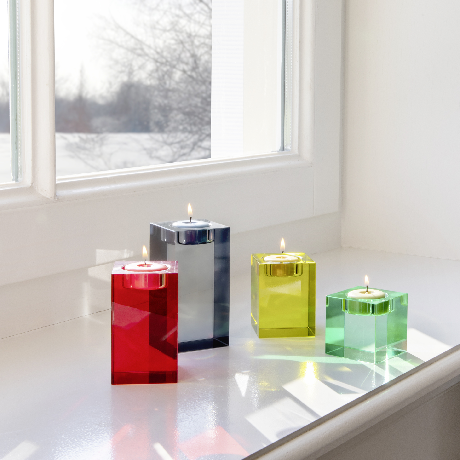 Vier farbige Glasblock-Kerzenhalter (rot, grau, gelb, grün) mit brennenden Teelichtern auf einer Fensterbank vor verschneiter Landschaft.