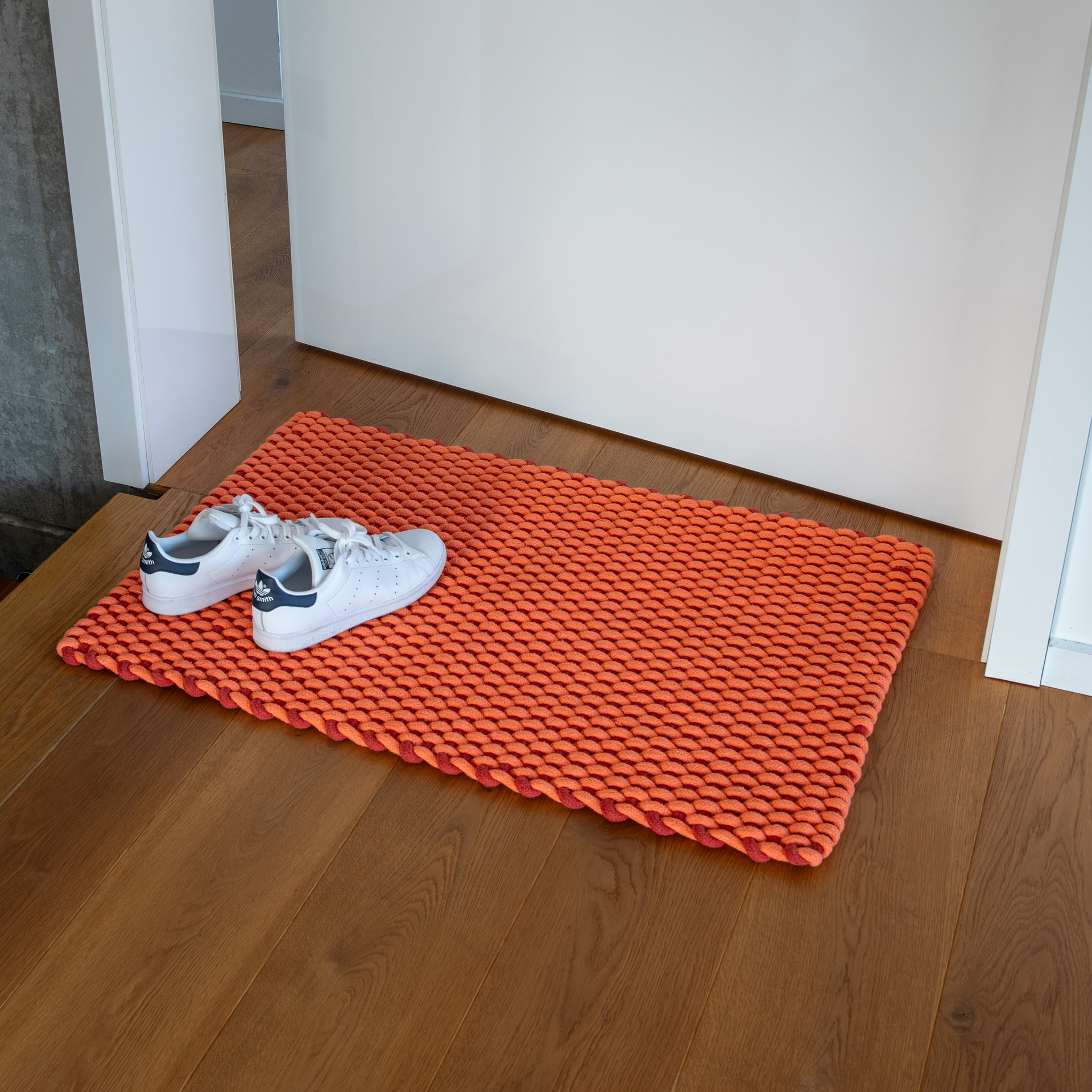 Handgewebte Fußmatte 'Lida' in Coral. Das geometrische Webmuster besteht aus harmonisch abgestimmten Koralle und Rotorange Tönen. Rechteckige Form.