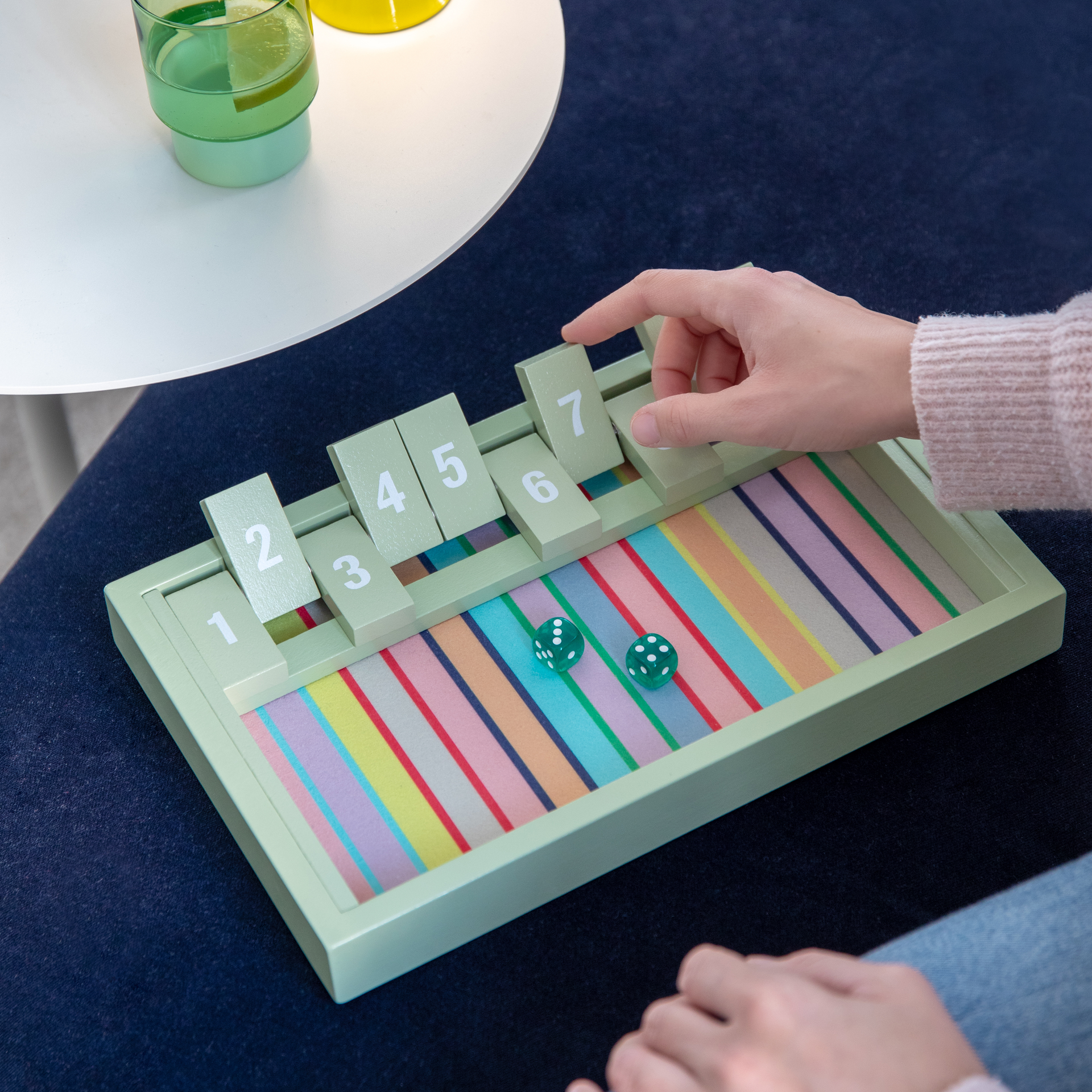 Das Holzspiel „Shut the Box“ mit Klappen 1-10, zwei grüne Würfel liegen in der Box auf bunten Streifen.