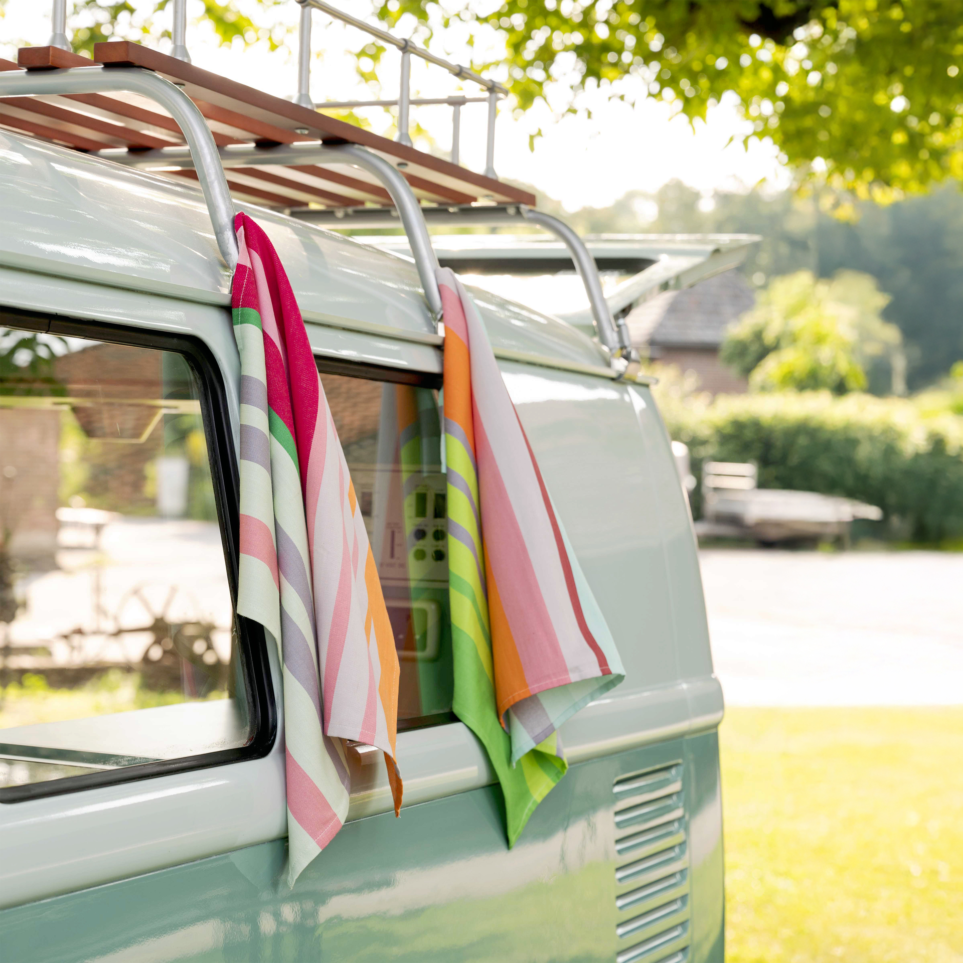 Zwei gestreifte Trockentücher hängen an einem mintgrünen VW Bus mit Dachgepäckträger. Die Tücher sind farbenfroh mit Streifen in Pink, Grün, Orange und Grau. Im Hintergrund ist eine grüne Wiese.