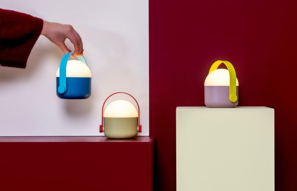 Drei farbenfrohe, tragbare Laternen mit leuchtendem Licht vor zweifarbigem Hintergrund. Eine Hand hält oben links die blaue Lampe. Die mintgrüne steht auf dunkelroter Stufe, die lila leuchtet auf cremefarbenem Podest.