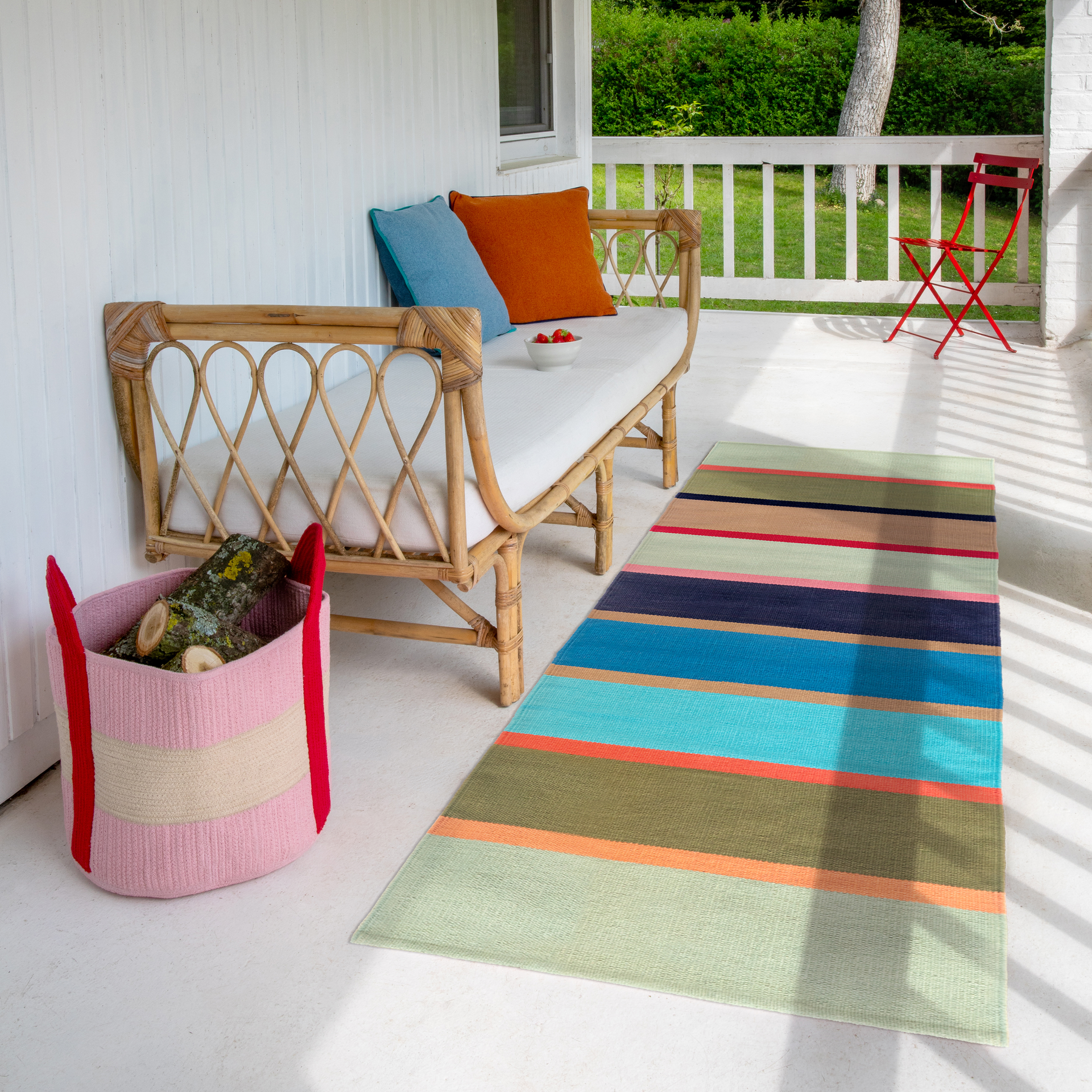 Veranda mit Bambussofa, weißen Polstern und bunten Kissen; Schale Erdbeeren. Links Korb mit Holzscheiten. Bunter Streifenteppich, Sonnenstreifen, hinten roter Klappstuhl.