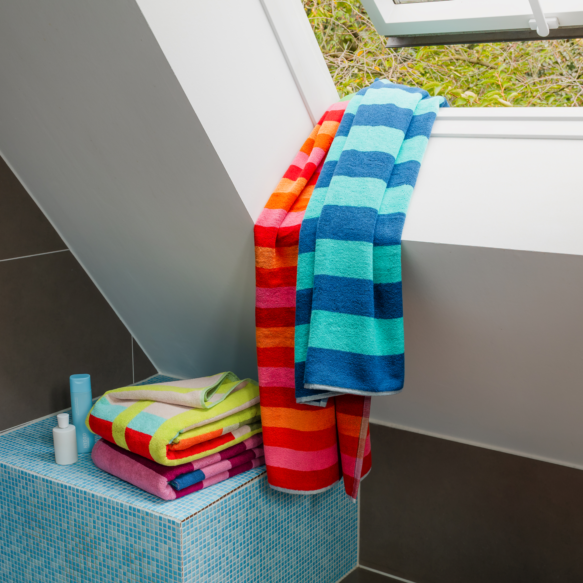 Serviette de bain Milo pliée verticalement, à larges rayures horizontales. En haut, bleu clair et foncé ; au milieu, vert clair et foncé. En bas, rayures vives rose, orange et rouge. Coton doux.