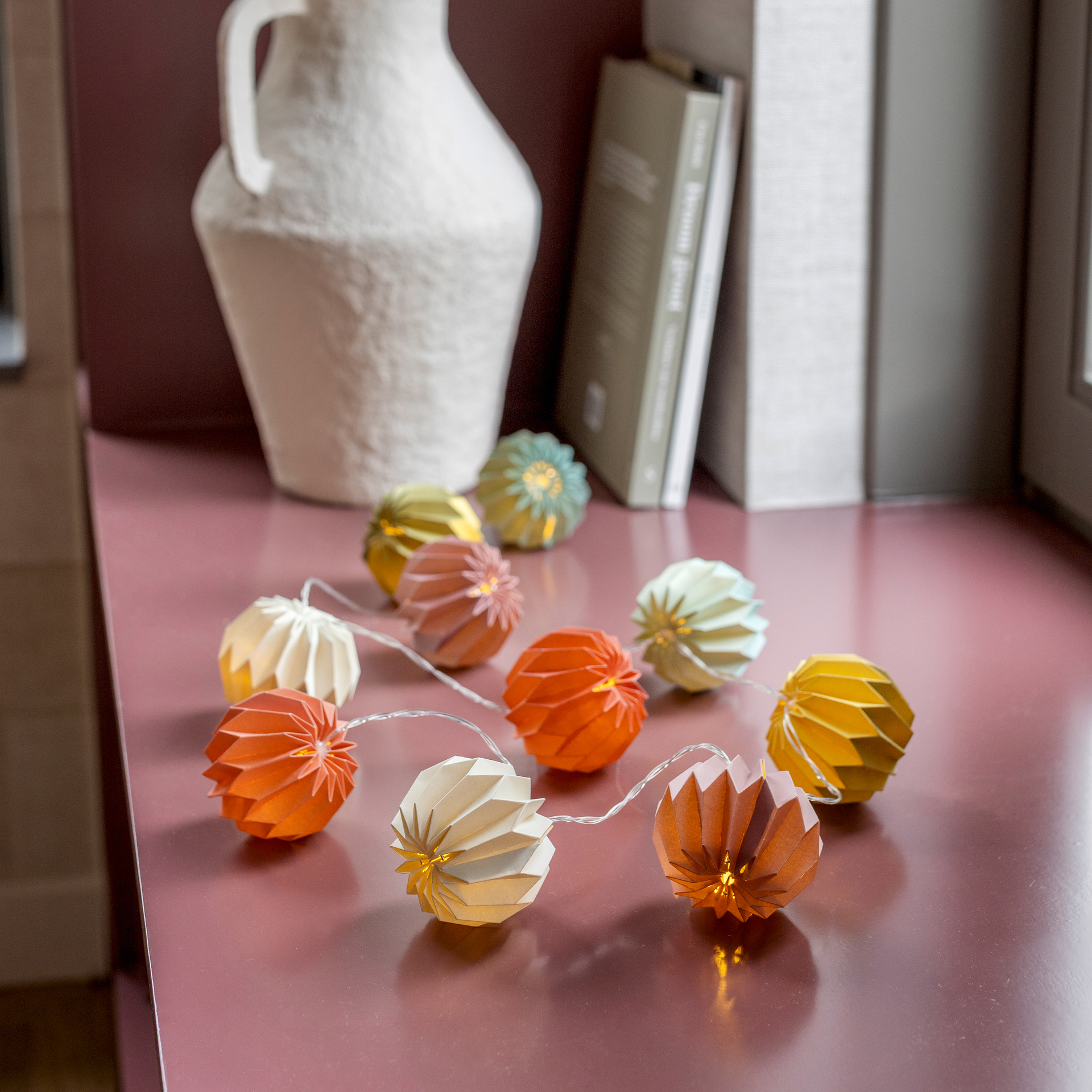 Auf einer rosa Oberfläche liegt die Papier-Lichterkette Yazz mit bunten, leuchtenden Papierornamenten. Im Hintergrund eine weiße Vase und Bücher.