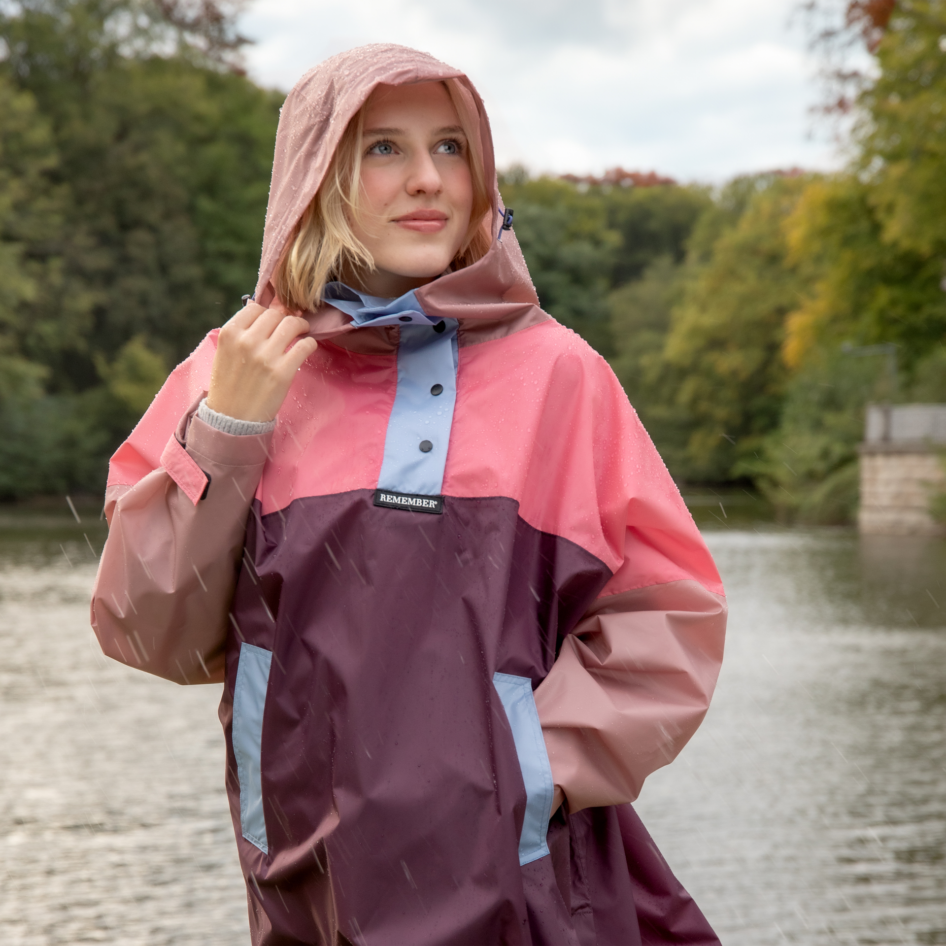 Freigestelltes, farbenfrohes Regencape Ida mit Kapuze und Druckknöpfen, in Pink-, Braun- und Blautönen. Seitliche Eingriffstaschen sind erkennbar.