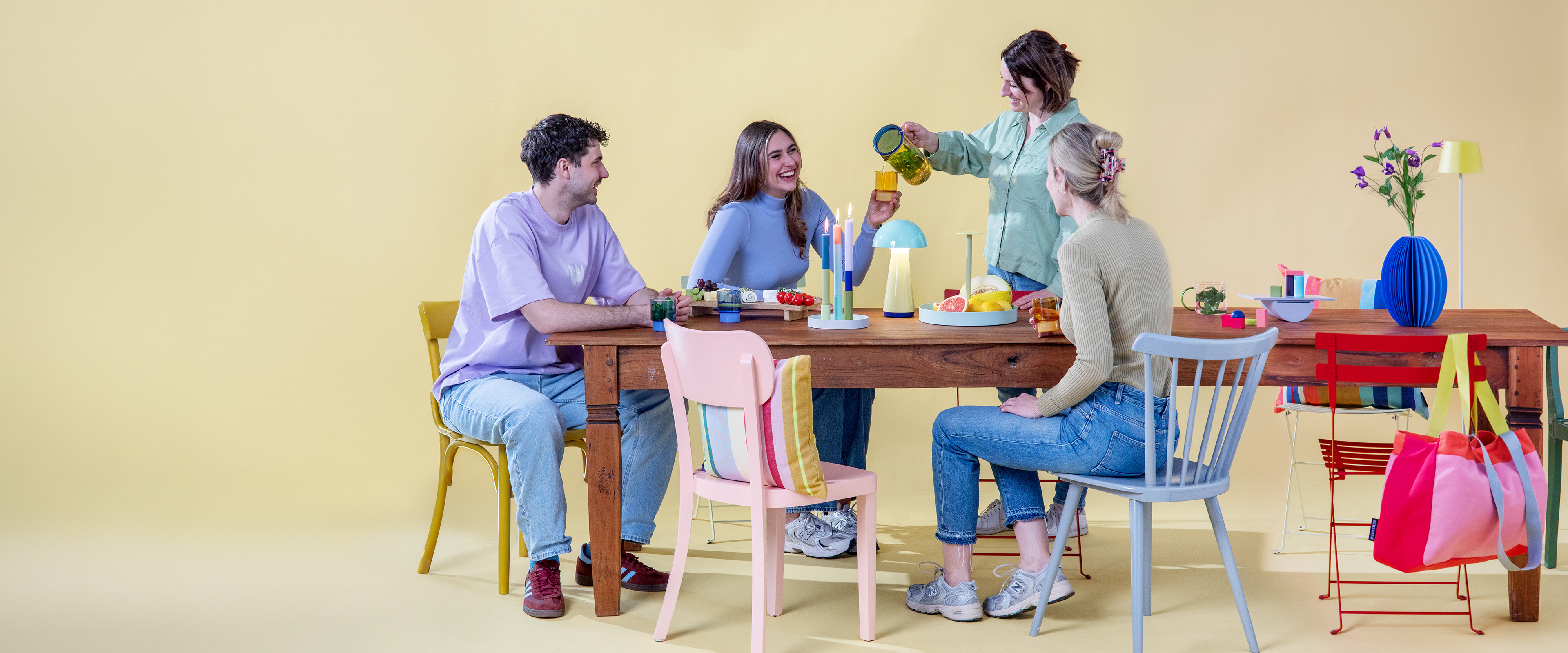 Vier junge Leute sitzen an einem Holztisch mit farbenfrohen Stühlen vor gelbem Hintergrund. Eine Frau schenkt Getränke ein, während die anderen lachen. Obst, Kerzen und eine Lampe stehen auf dem Tisch.