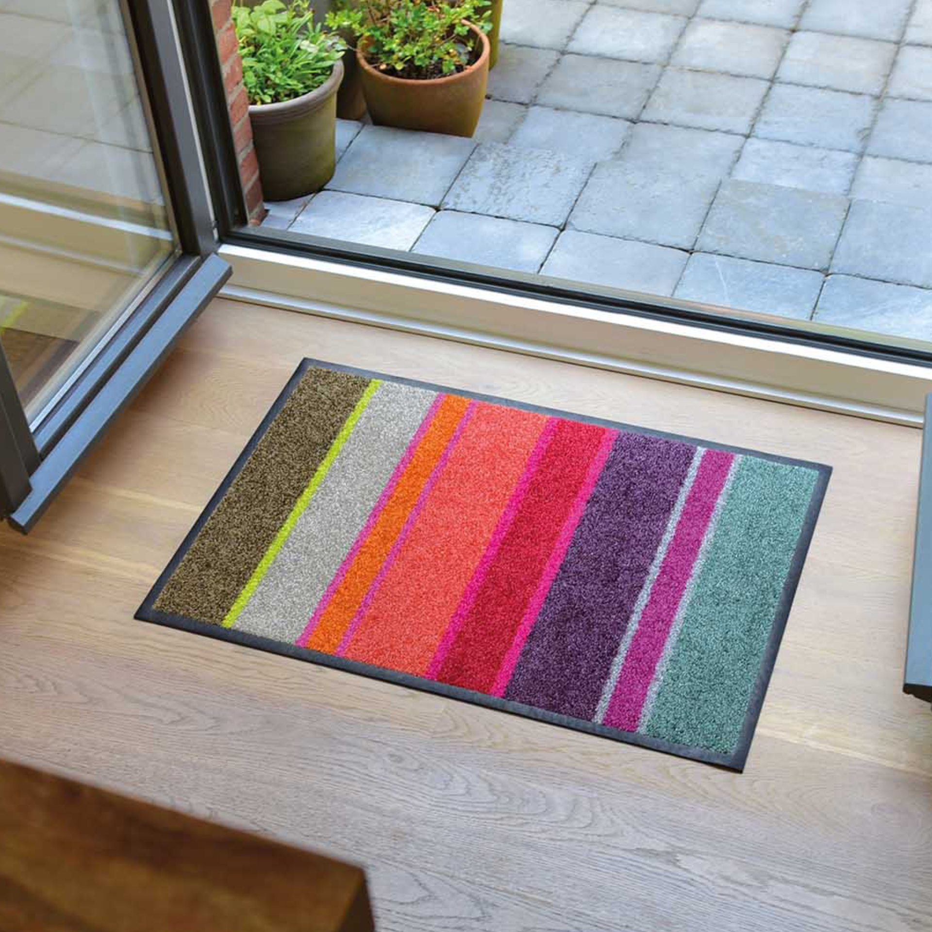 Rechteckige Fußmatte 'Largo' mit einem schwarzen Rand. Das Muster besteht aus vertikalen Streifen in Grün, Pink, Lila, Rot, Orange, Beige und Braun.
