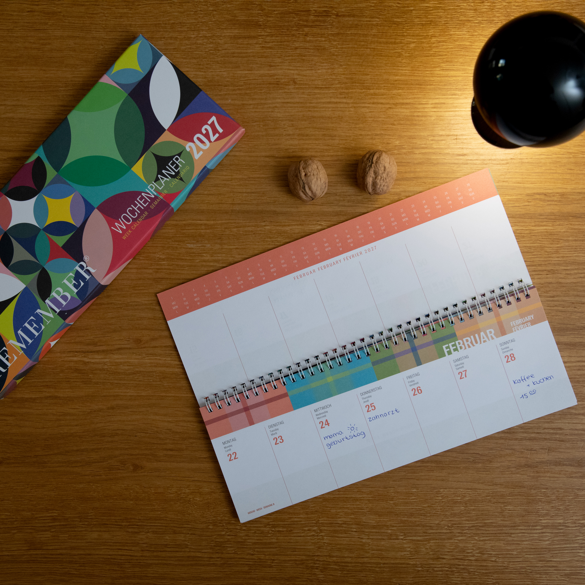 Calendario settimanale aperto di febbraio 2027 su scrivania di legno con appunti «Compleanno della mamma» e «Caffè + torta». A sinistra, confezione «Agenda settimanale 2027». Visibili due noci e una lampada nera.