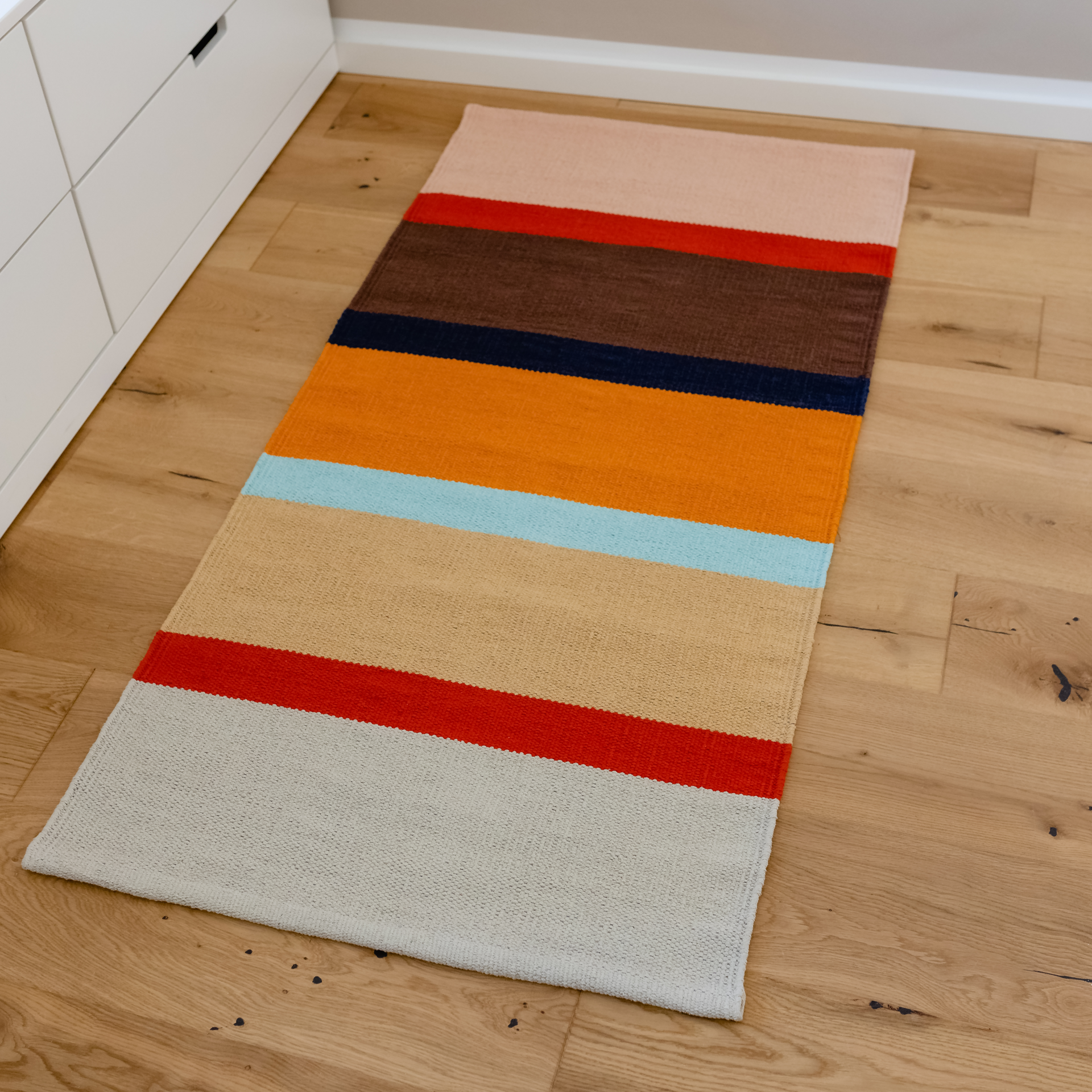 Tapis de sol 'Zaza' à poils courts, tissé à la main, en coton, avec des rayures colorées dans les tons marron, rouge, orange et bleu sur un fond clair.