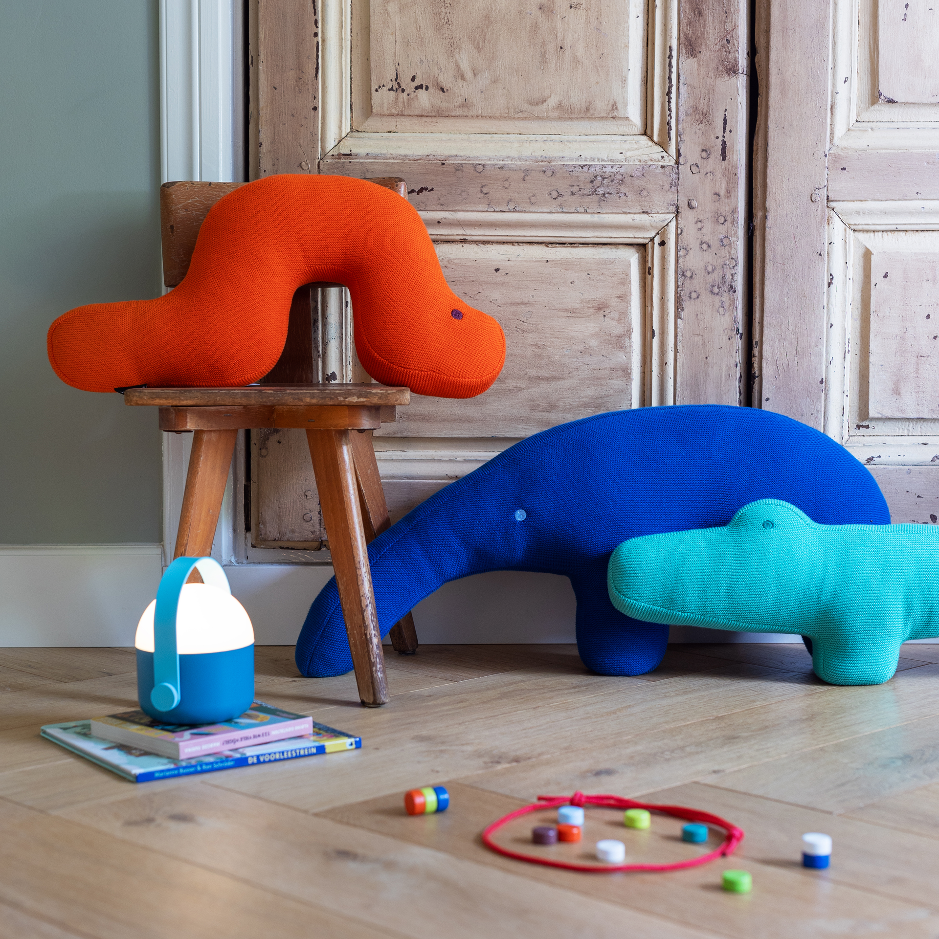 Produktdarstellung mit Kuschelkissen: Orangefarbener Wurm auf Stuhl, blauer Wal, türkises Krokodil („Kroko Karl“). Spielzeug und Lampe im Vordergrund.
