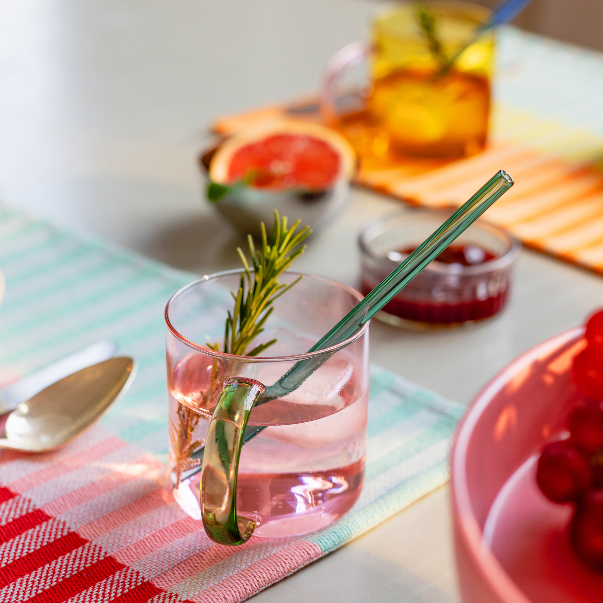 Ein rosefarbener Glasbecher 'Fred' mit grünem Henkel und Strohhalm, gefüllt mit einem Getränk und einem Zweig Rosmarin, steht auf einem Tisch mit Speisen.