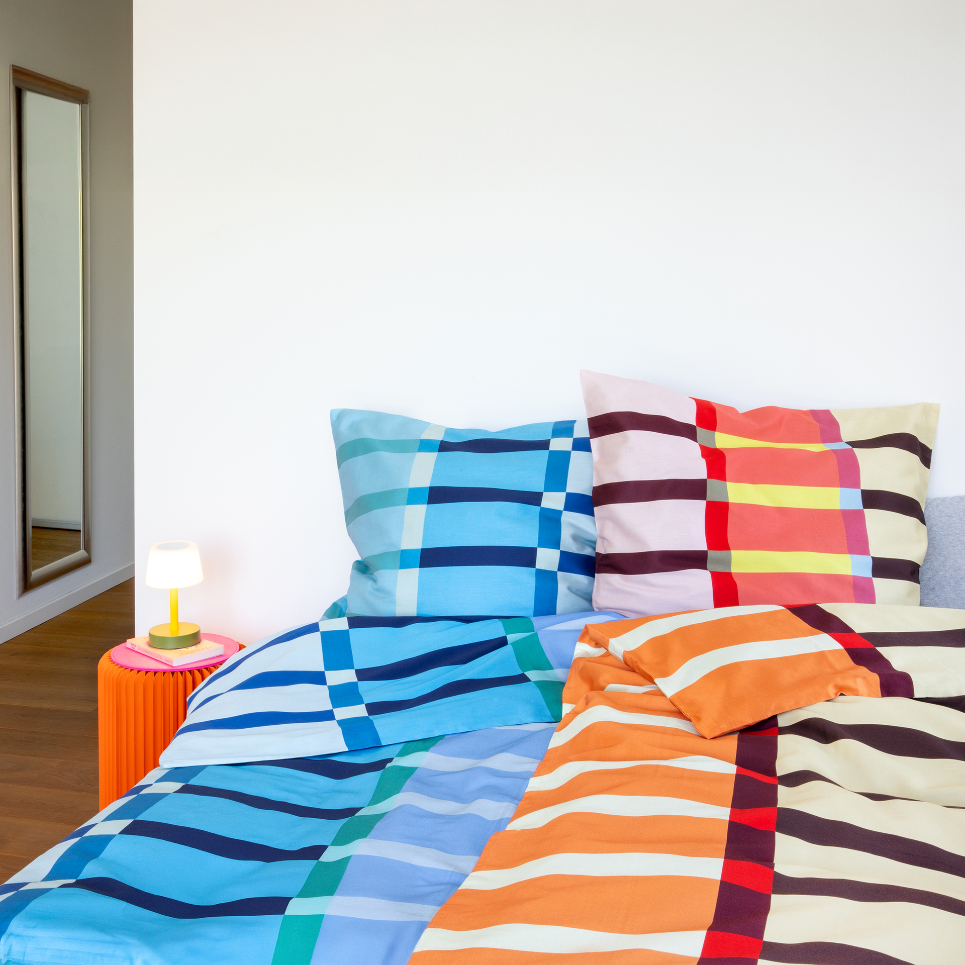 Bettwäsche 'Bari' im Detail: Bunt gestreifte und karierte Kissen und Decke auf einem Bett. Im Hintergrund ein Spiegel und ein Nachttisch mit Lampe.
