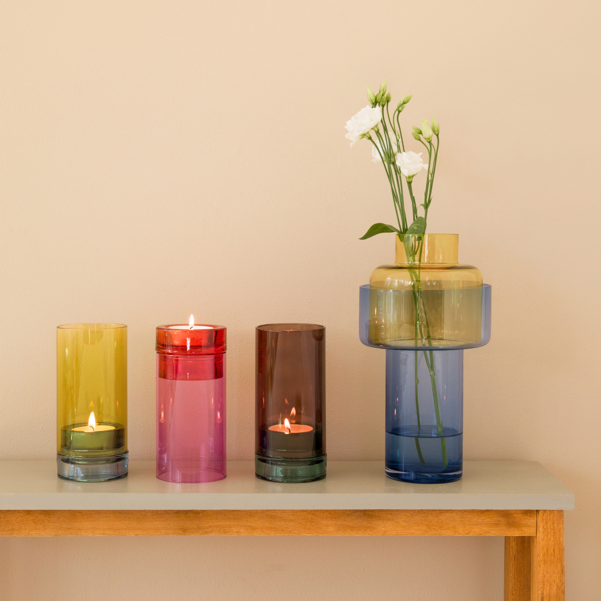 Vier Glasobjekte auf einer Tischplatte. Von links: gelbes Windlicht mit Teelicht, pinkes Windlicht, braunes Windlicht mit Teelicht, blau-gelbe Vase mit weißen Blumen.