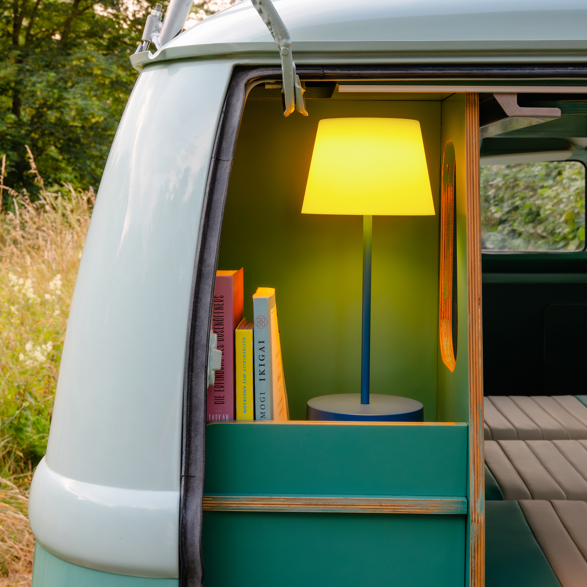 Leuchtende 'midnight' Tischleuchte Oscar in einem Campingbus. Bücher stehen daneben. Warmes Licht erhellt den Innenraum.