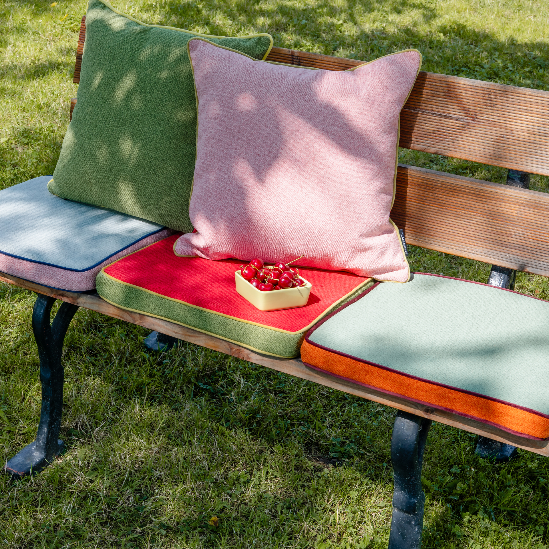 Auf einer Holzbank liegen Kissen in Grün, Rosa, Rot und Mint. Eine Schale mit Kirschen steht darauf. Die Kissen haben farblich abgesetzte Paspeln. Rasen ist im Hintergrund.