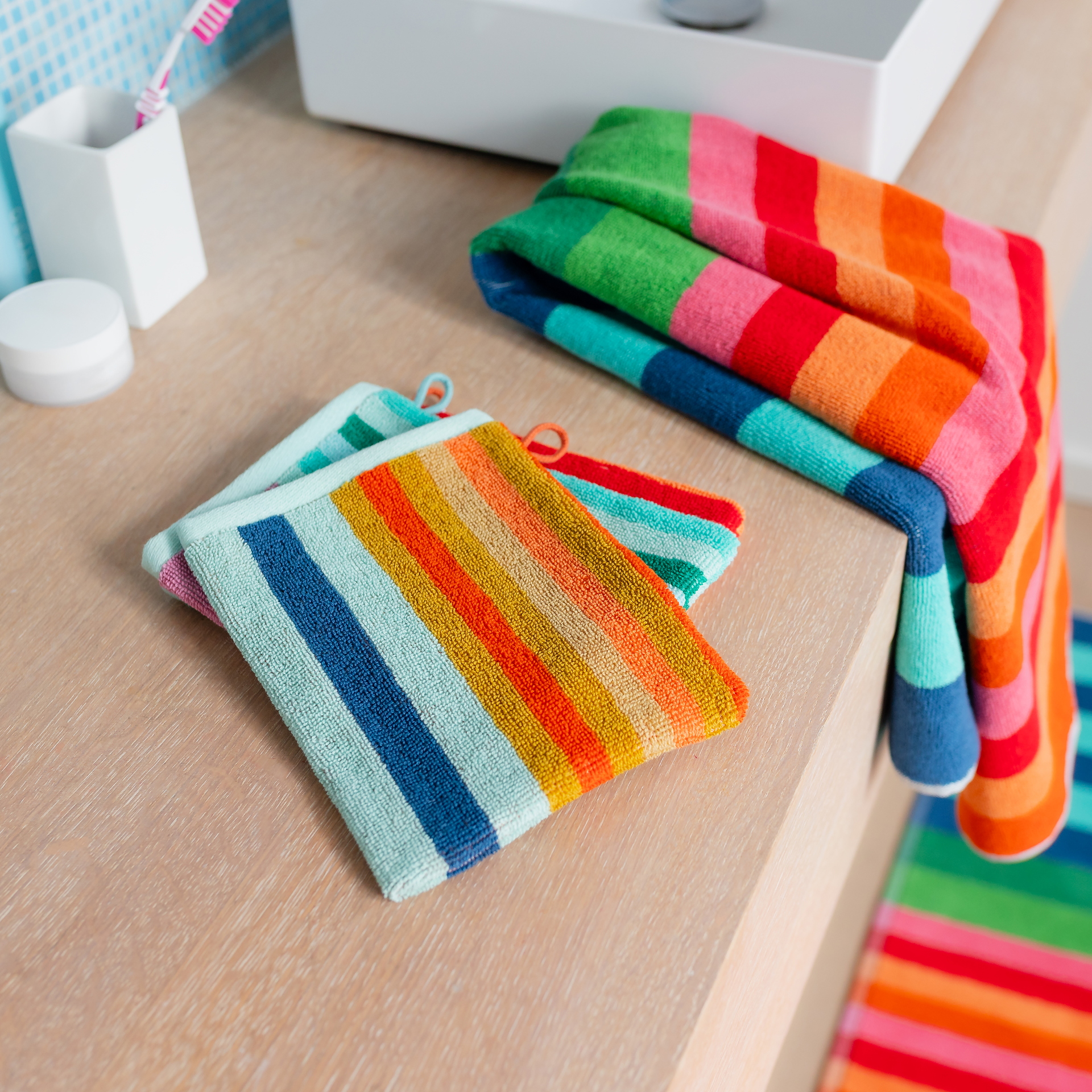 Lot de 3 gants de toilette Milo colorés et rayés sur fond blanc. Chaque gant, doux pour la peau, a un bord vert menthe avec boucle de suspension. Rayures verticales en tons chauds, terreux et froids.