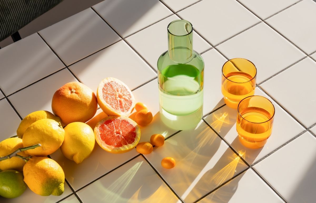 Zitrusfrüchte auf weißem Fliesenboden: ganze und halbierte Grapefruits mit rotem Fruchtfleisch, Zitronen, Kumquats und eine Limette. Daneben eine grüne Glaskaraffe und zwei orange Gläser, im starken Sonnenlicht mit langen Schatten.