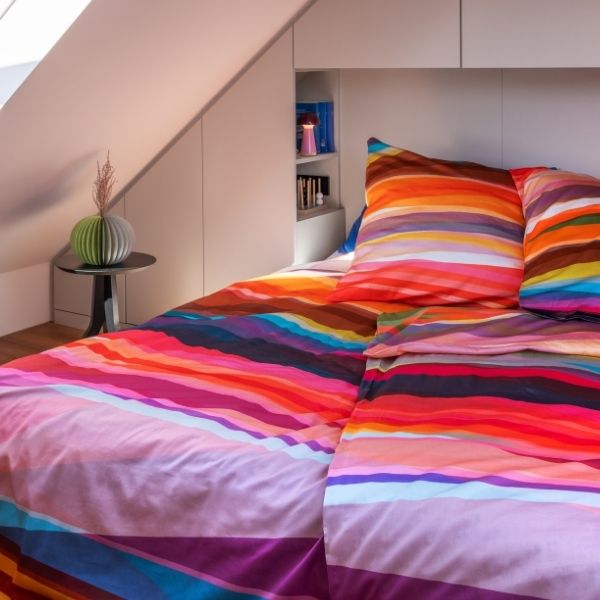 Chambre confortable sous les combles : lit avec draps à rayures vives (rouge, orange, rose, violet, bleu). À gauche, petite table d'appoint avec vase vert moderne. Derrière, placards encastrés blancs avec étagère.