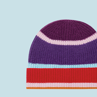 Detailaufnahme einer gestreiften, gestrickten Mütze in Lila, Pink, Rot, Orange und Hellblau vor hellblauem Hintergrund.