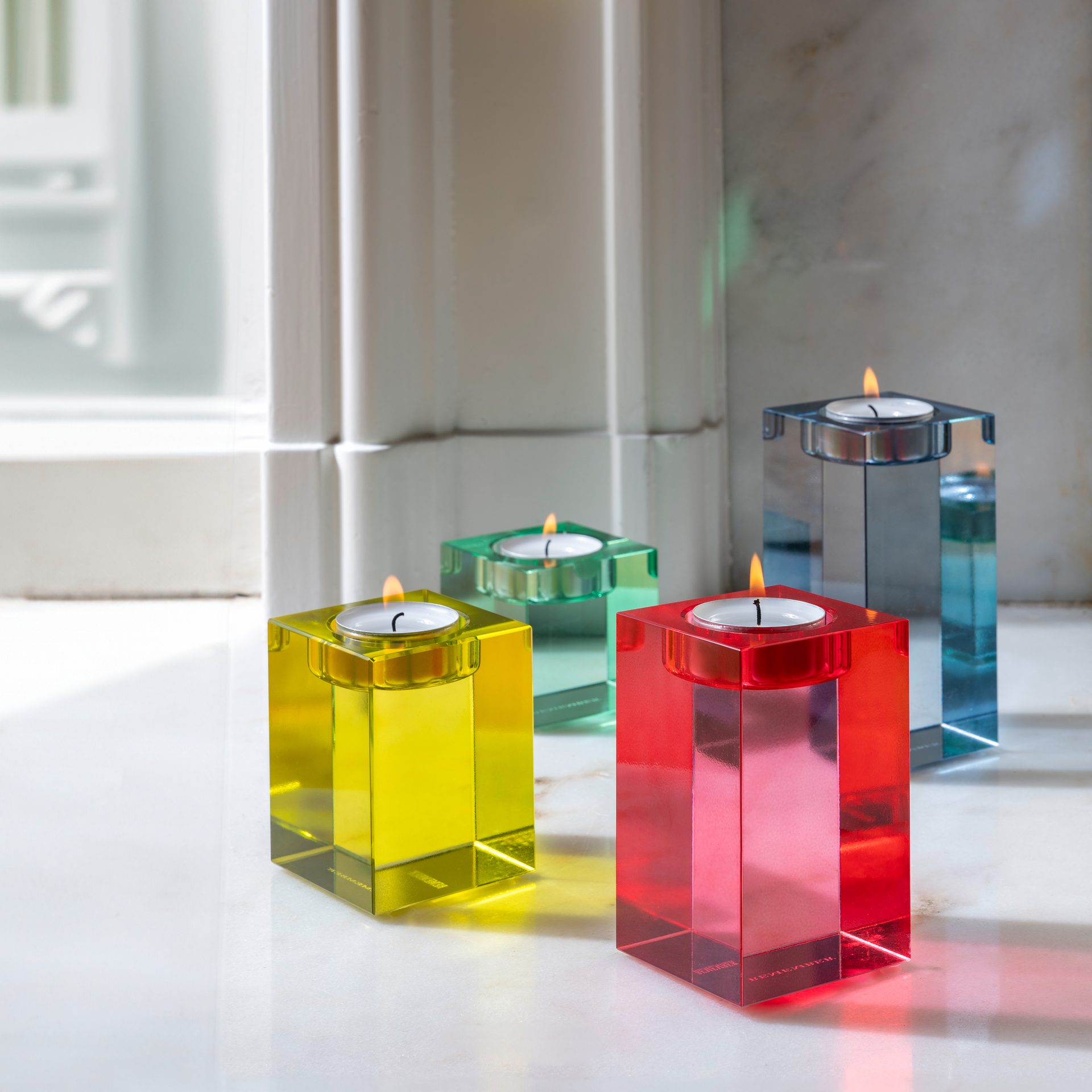 Vier farbige Glasblock-Kerzenhalter 'Vela' in Rot, Gelb, Grün und Grau. Die Kerzenhalter sind quaderförmig mit Aussparungen für Teelichter.