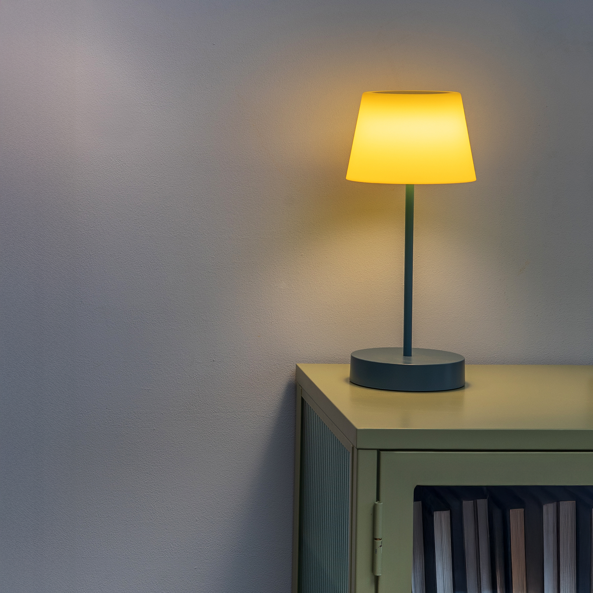 Auf einem Schrank steht ein angeschaltetes, hellgelb leuchtendes Tischleuchten-Modell „Oscar Midnight“ mit einem schlanken, dunkelgrauen Fuß und Lampenschirm.