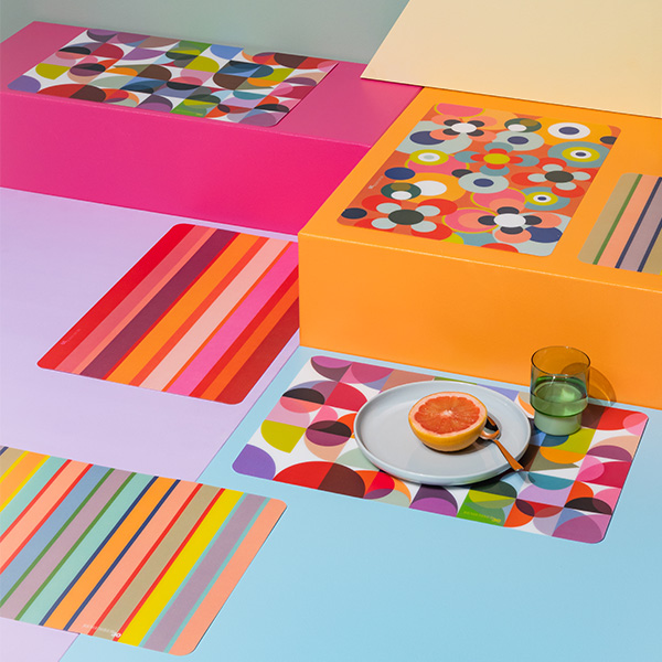 Mehrere farbenfrohe Tischsets auf bunten Quadern, darunter ein Teller mit Grapefruit und ein Glas Wasser. Die Tischsets sind geometrisch gemustert.