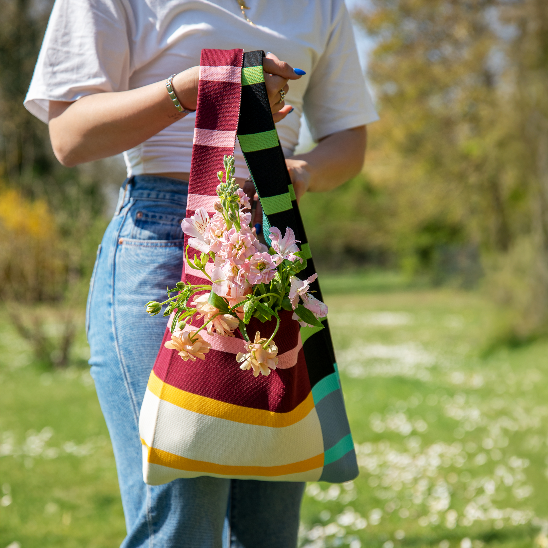 Detailaufnahme einer Person mit einer gestreiften Stricktasche mit Blumen in der Hand. Die Farben der Tasche sind Rot, Schwarz, Grün und Gelb.