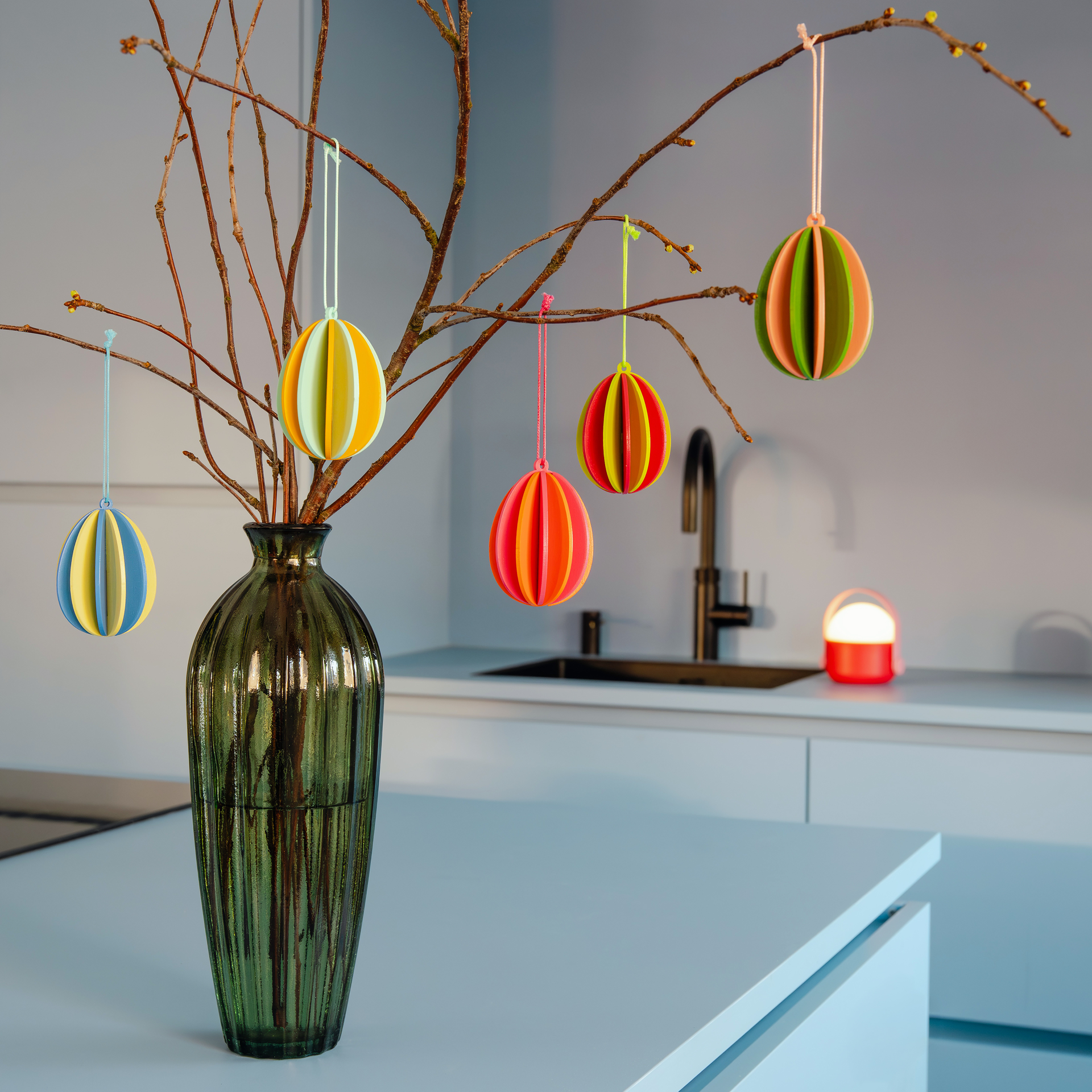 Ein Bund kahler Äste in einer grünen Glasvase steht auf einer Küchentheke. An den Ästen hängen bunte Papier-Ostereier. Im Hintergrund eine Spüle und eine rote Lampe.