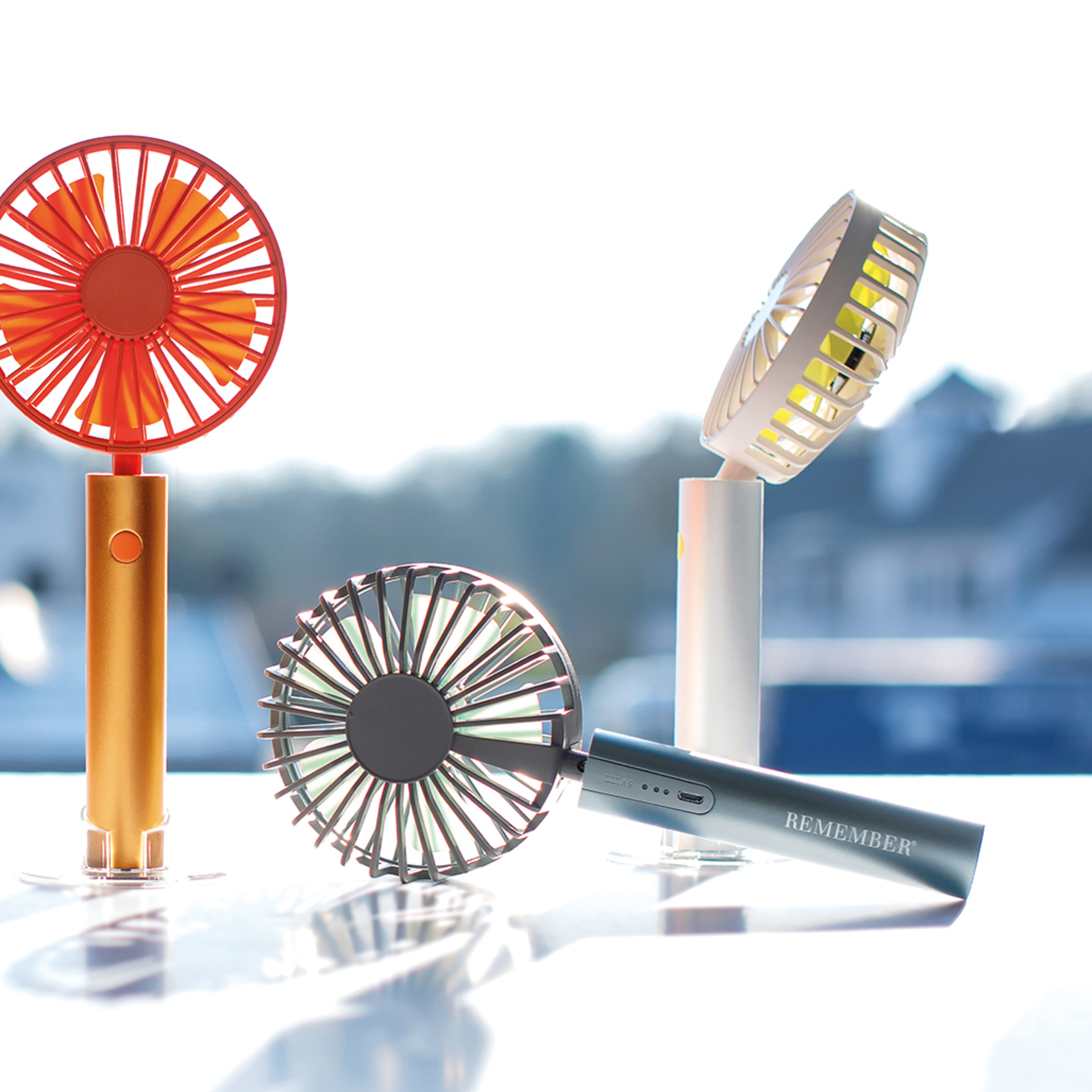 Vier Akku-Ventilatoren in Silber, Orange und Weiß mit transparenten Standfüßen. Ein Ventilator liegt, die anderen stehen.