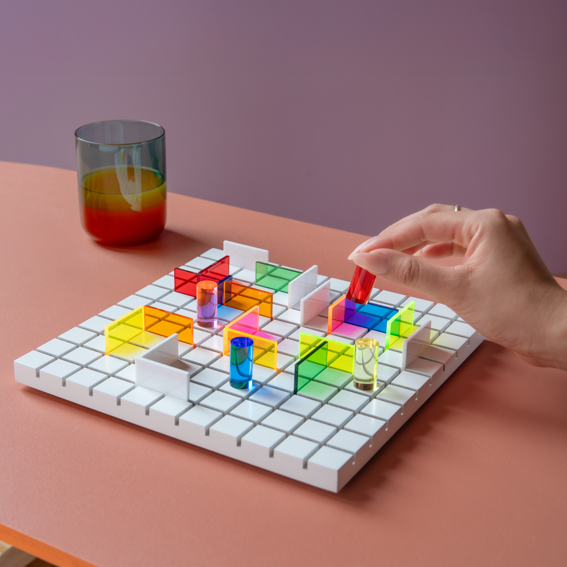 Ein Spielbrett mit bunten, durchsichtigen Steinen und einer Hand, die einen roten Stein platziert. Im Hintergrund ein Glas mit mehrfarbiger Flüssigkeit.