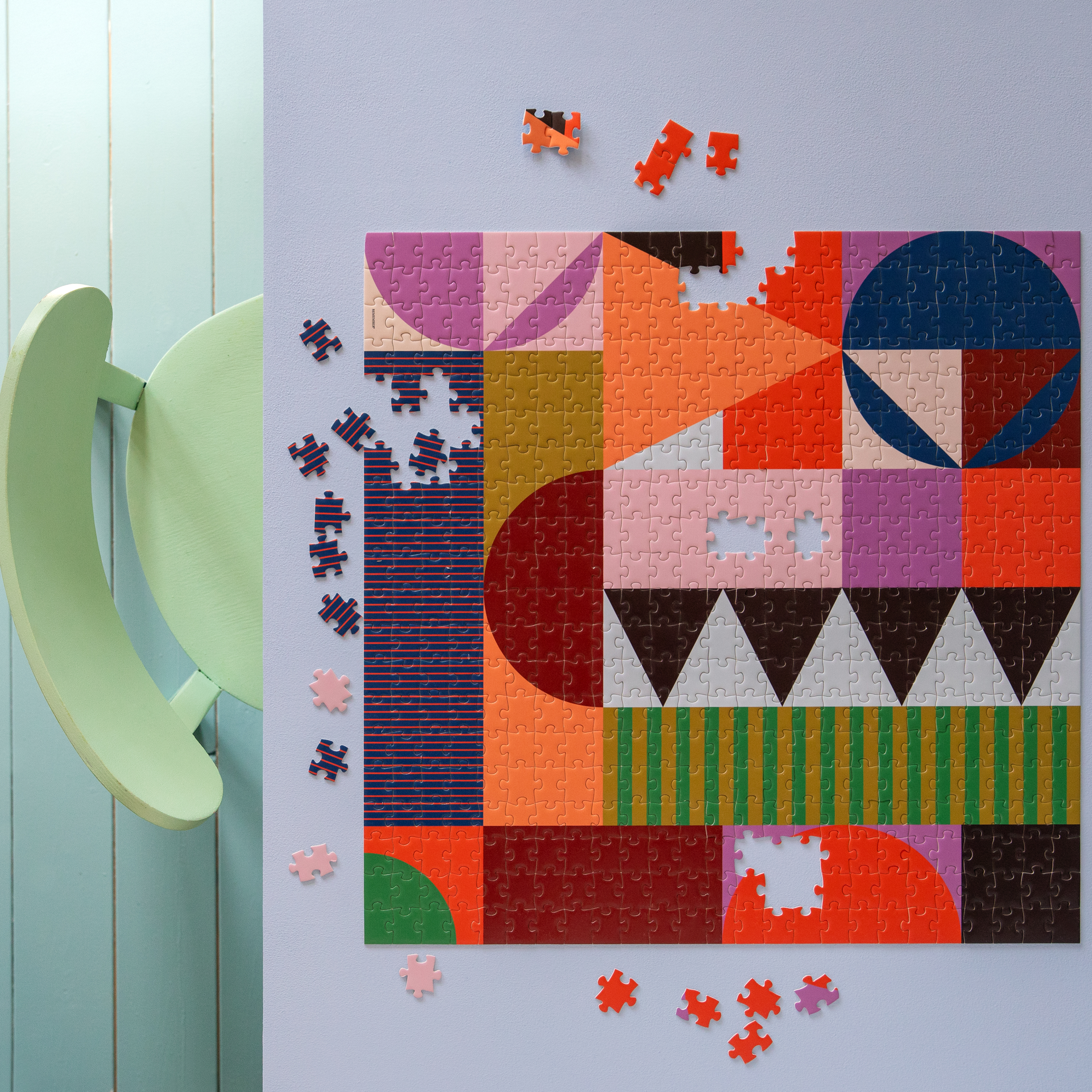 Ein fast fertiges, buntes Puzzle mit geometrischen Formen liegt auf einem Tisch, einige Puzzleteile liegen daneben. Im Hintergrund ist ein hellgrüner Stuhl zu sehen.