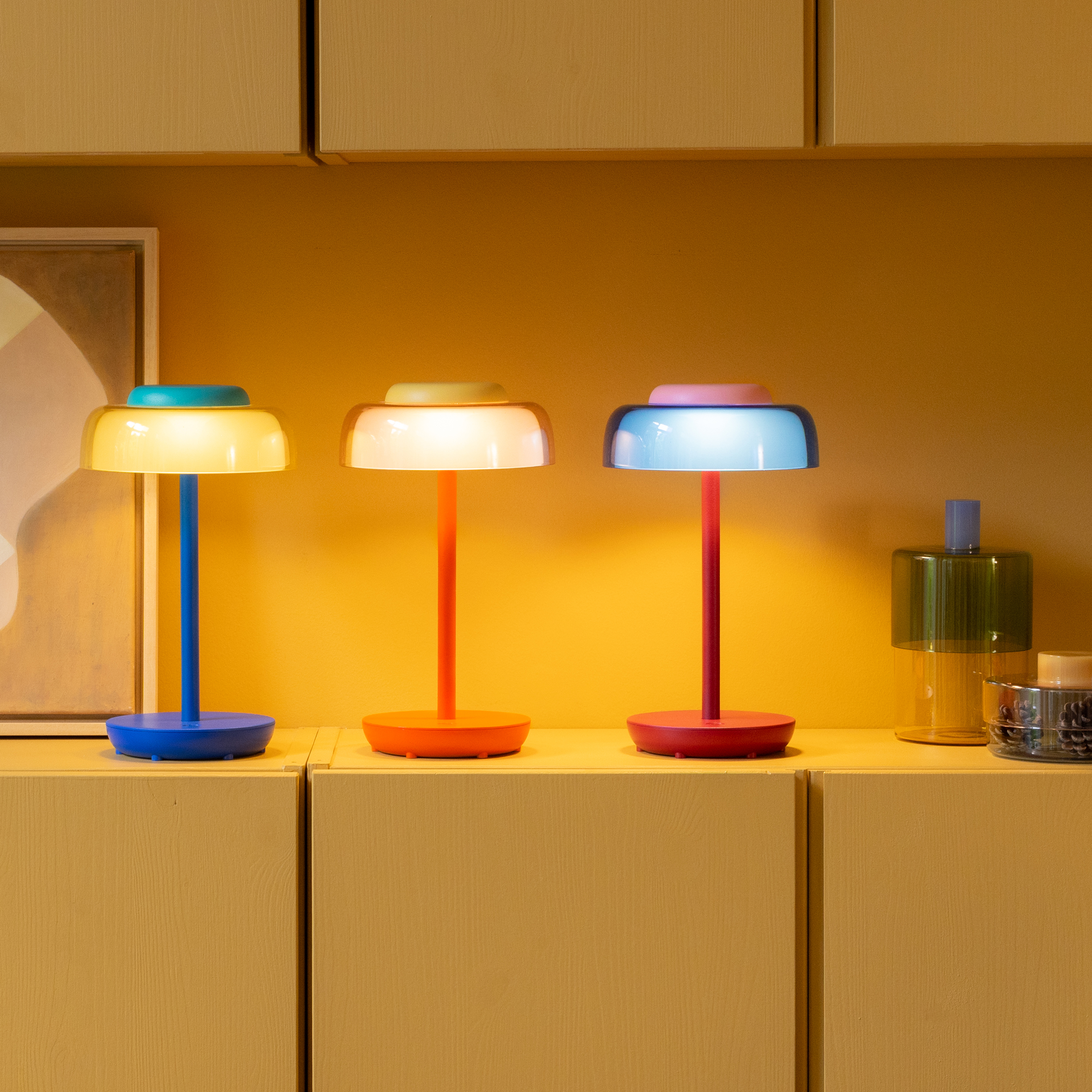 Tre lampade da tavolo Juno in vari colori su uno scaffale: blu con paralume giallo e cappuccio turchese, arancione brillante con dettagli gialli e rossa con paralume blu e cappuccio rosa. LED senza fili davanti a parete color miele.