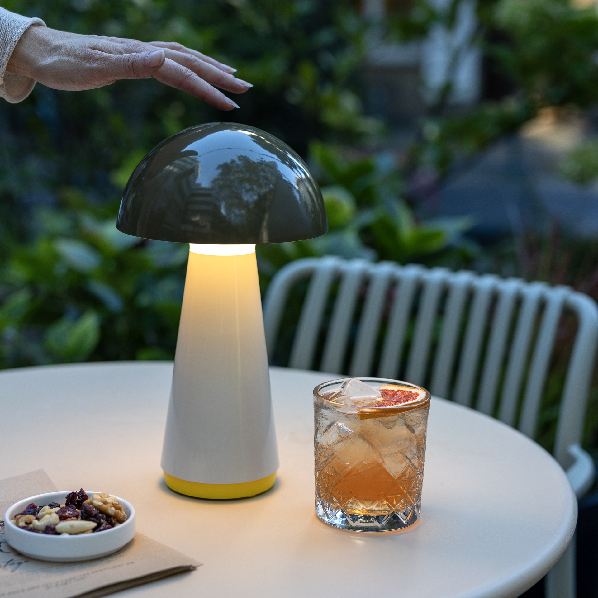 Eine olivenfarbene LED Tischleuchte im Pilzdesign steht beleuchtet auf einem Tisch. Eine Hand berührt den Lampenschirm. Daneben ein Cocktail und eine Schale mit Nüssen.
