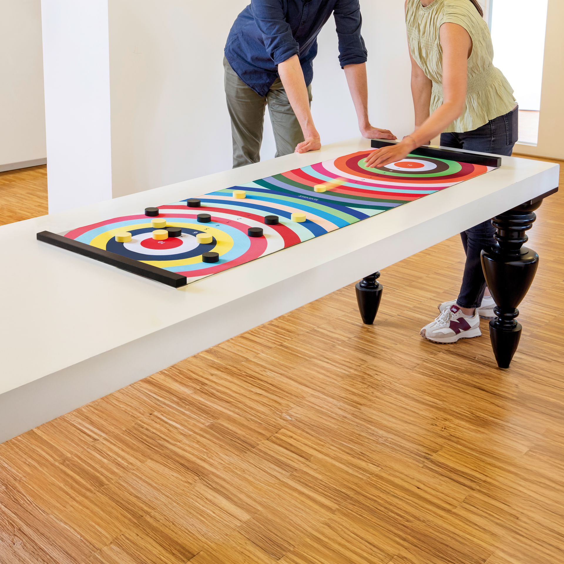 Zwei Personen spielen Colour Curling auf einem Tisch. Das Spielfeld mit farbigen Zielkreisen ist mit schwarzen und gelben Spielsteinen versehen.
