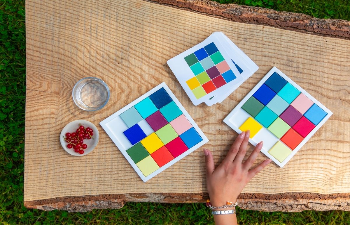 Nahaufnahme: zwei weiße Puzzletabletts mit bunten Quadraten auf rustikalem Holz. Eine Hand setzt einen gelben Stein ins rechte, fast volle Puzzle. Daneben Farbkarten, Wasser und Schale mit roten Johannisbeeren im Gras.