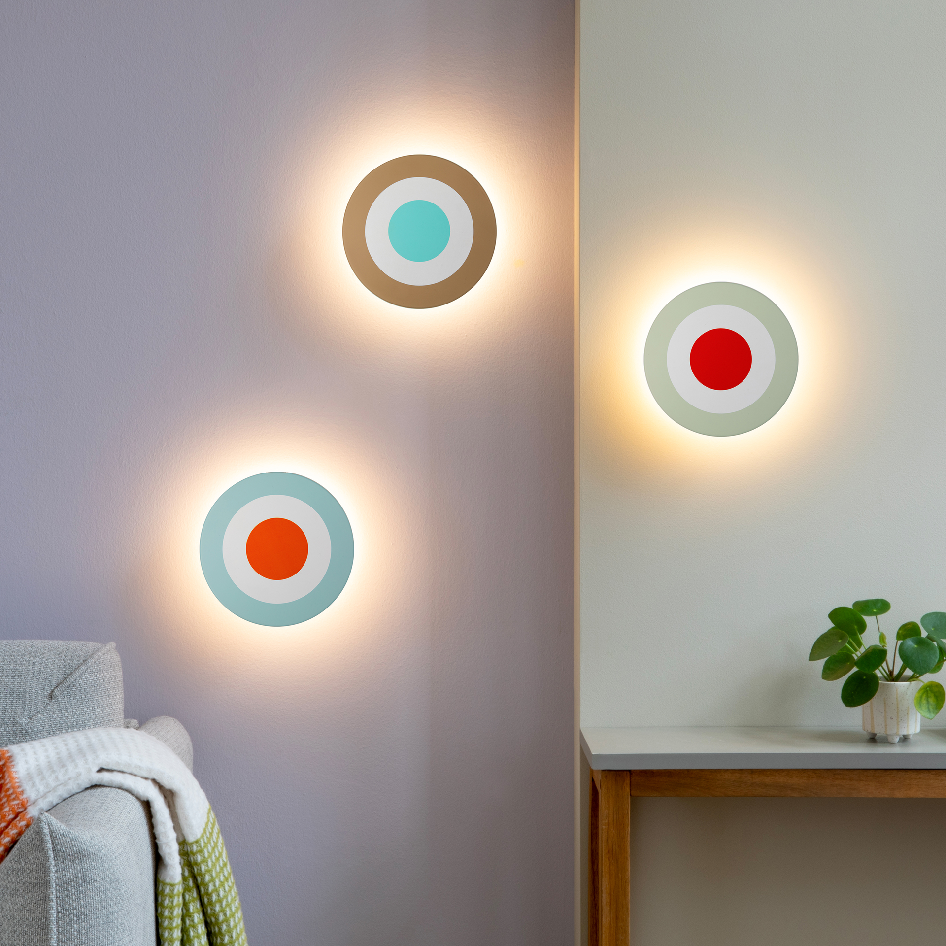 Trois appliques murales DOT à batterie diffusent une lumière blanc chaud sur deux murs. Chaque applique ronde, à cercles concentriques turquoise, orange et rouge, éclaire doucement le canapé et la console.