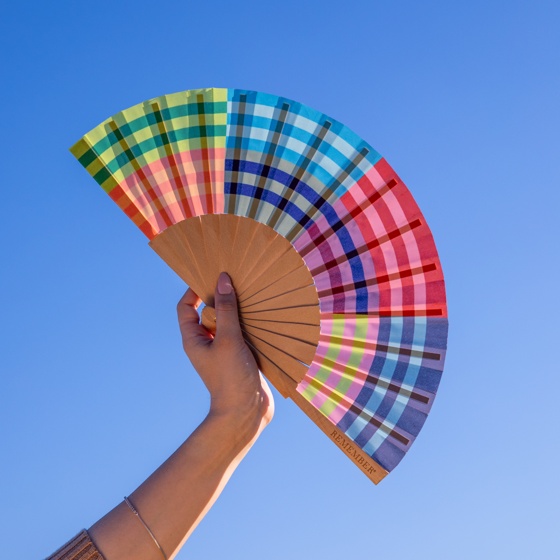 Eine Hand hält einen geöffneten, bunten Handfächer vor blauem Himmel. Der Fächer hat ein Karomuster in Regenbogenfarben und Holzstreben.
