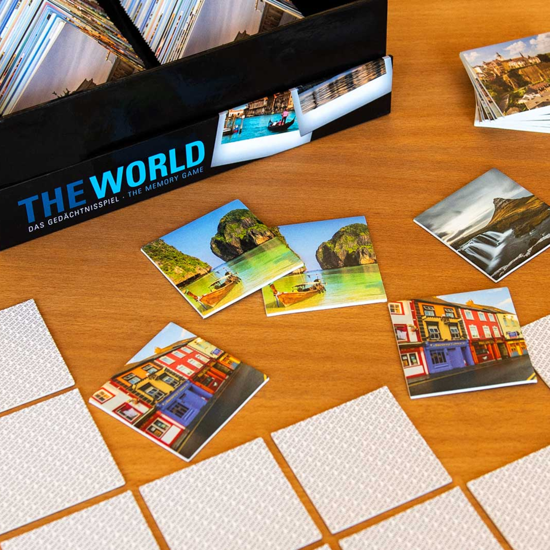 Das Memory-Spiel 'The World' liegt auf einem Holztisch. Gezeigt werden die Box, Kartenstapel und aufgedeckte Karten mit Motiven aus aller Welt.