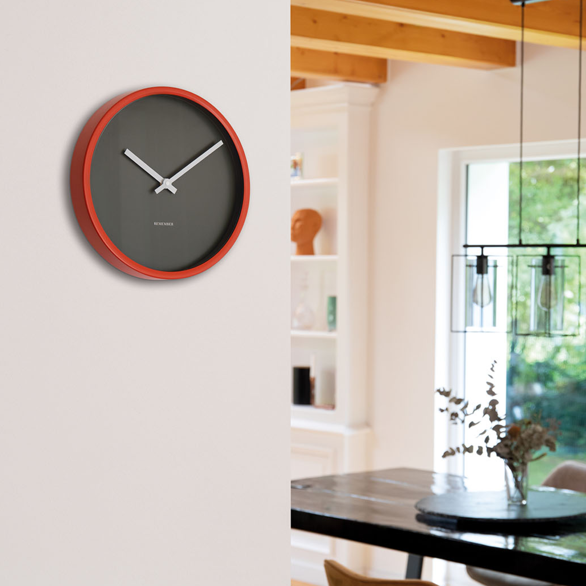 Wanduhr 'Mocca' an einer weißen Wand. Die Uhr hat ein rotes Gehäuse, graues Zifferblatt und weiße Zeiger. Im Hintergrund ist ein Esszimmer mit Tisch und Stühlen zu sehen.