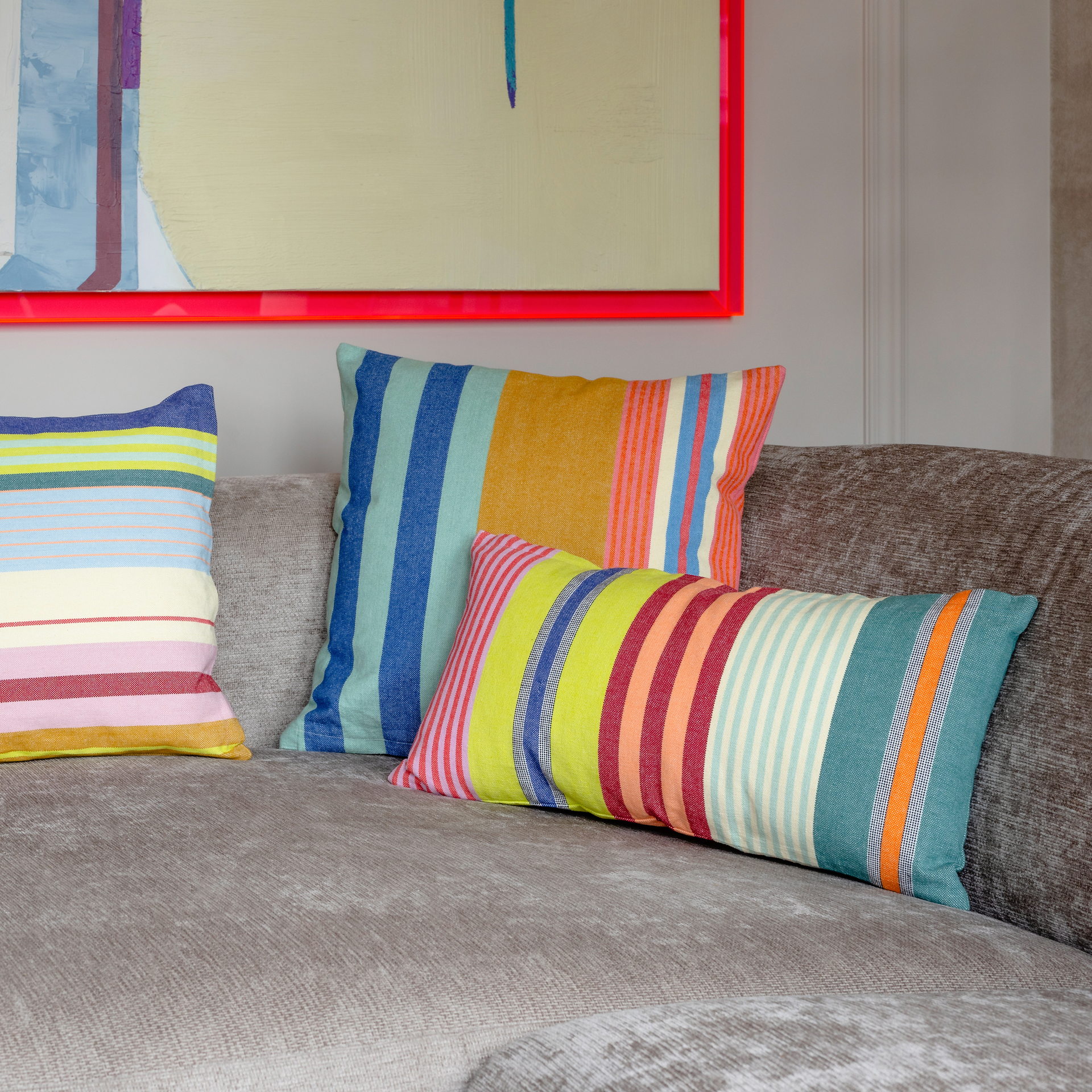 Drei Baumwoll-Kissen 'Barbados' mit bunten Streifen auf einem Sofa. Im Hintergrund hängt ein gerahmtes Bild mit hellen Farben.