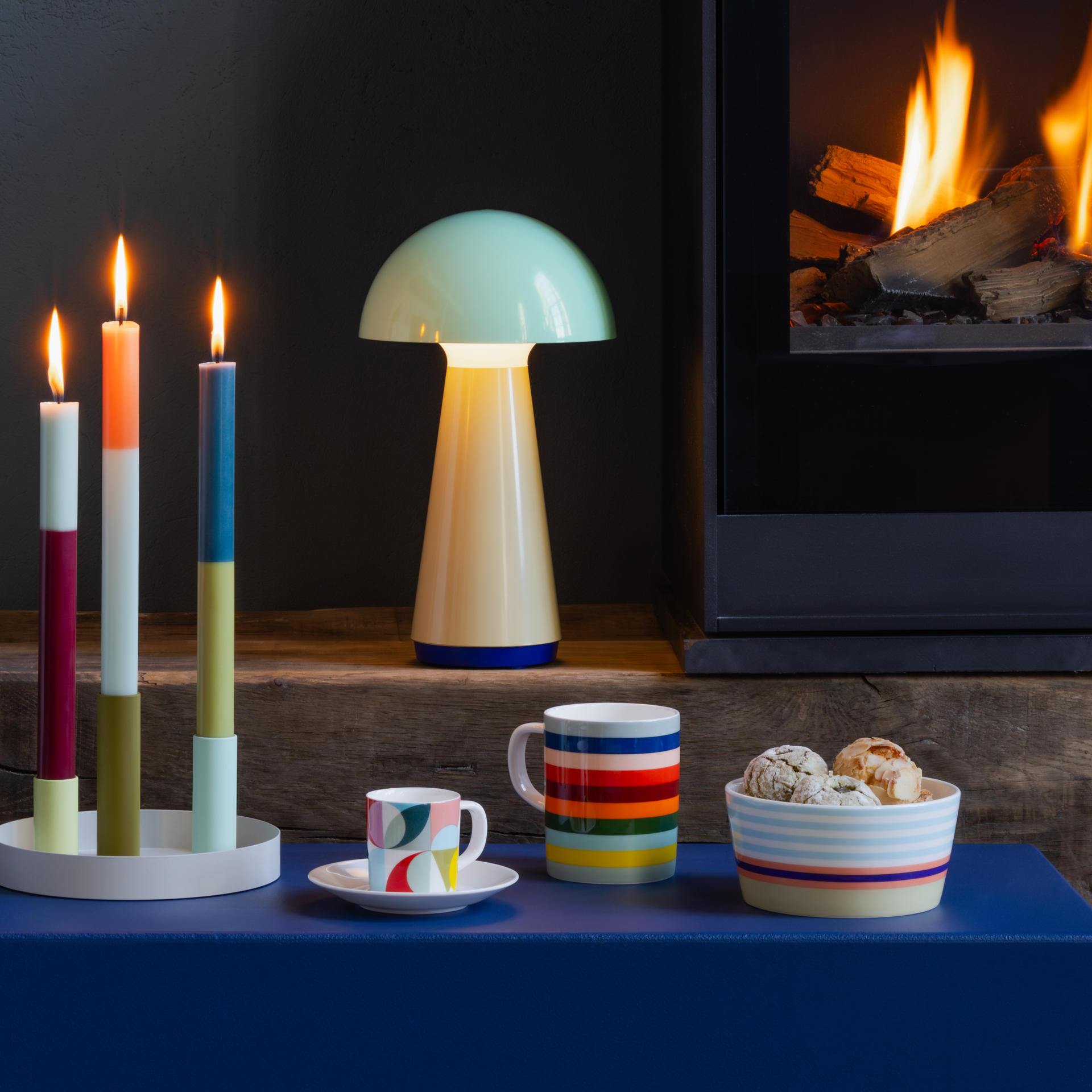 Ambiente mit brennendem Kamin, Pilzlampe und farbigen Kerzen. Vorne auf blauem Tisch eine 'Lorenzo' Tasse mit Streifen, eine kleine Espressotasse und eine Schale mit Keksen.