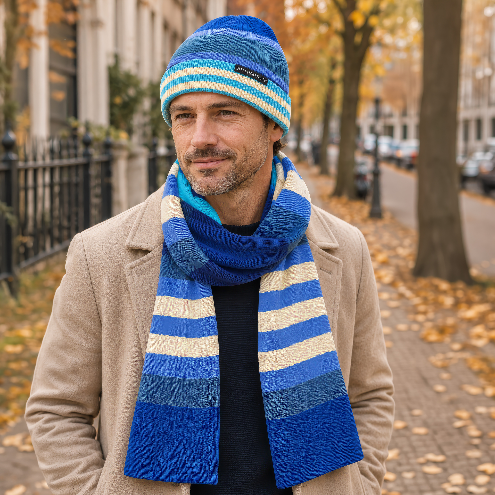 Bonnet en coton tricoté à rayures horizontales bleu et crème. Rayures larges sur le dessus, bleu foncé, moyen et clair ; rayures fines sur le bord rabattu, bleu clair et beige. Étiquette noire «REMEMBER®» devant.