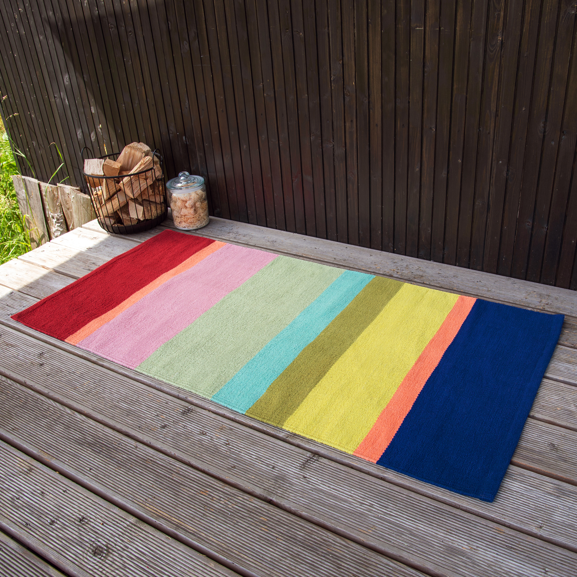 Un tapis de sol rayé 'Riva' en rouge, rose, vert, jaune et bleu est posé sur un sol en bois. En arrière-plan, on voit du bois et un panier de bois.