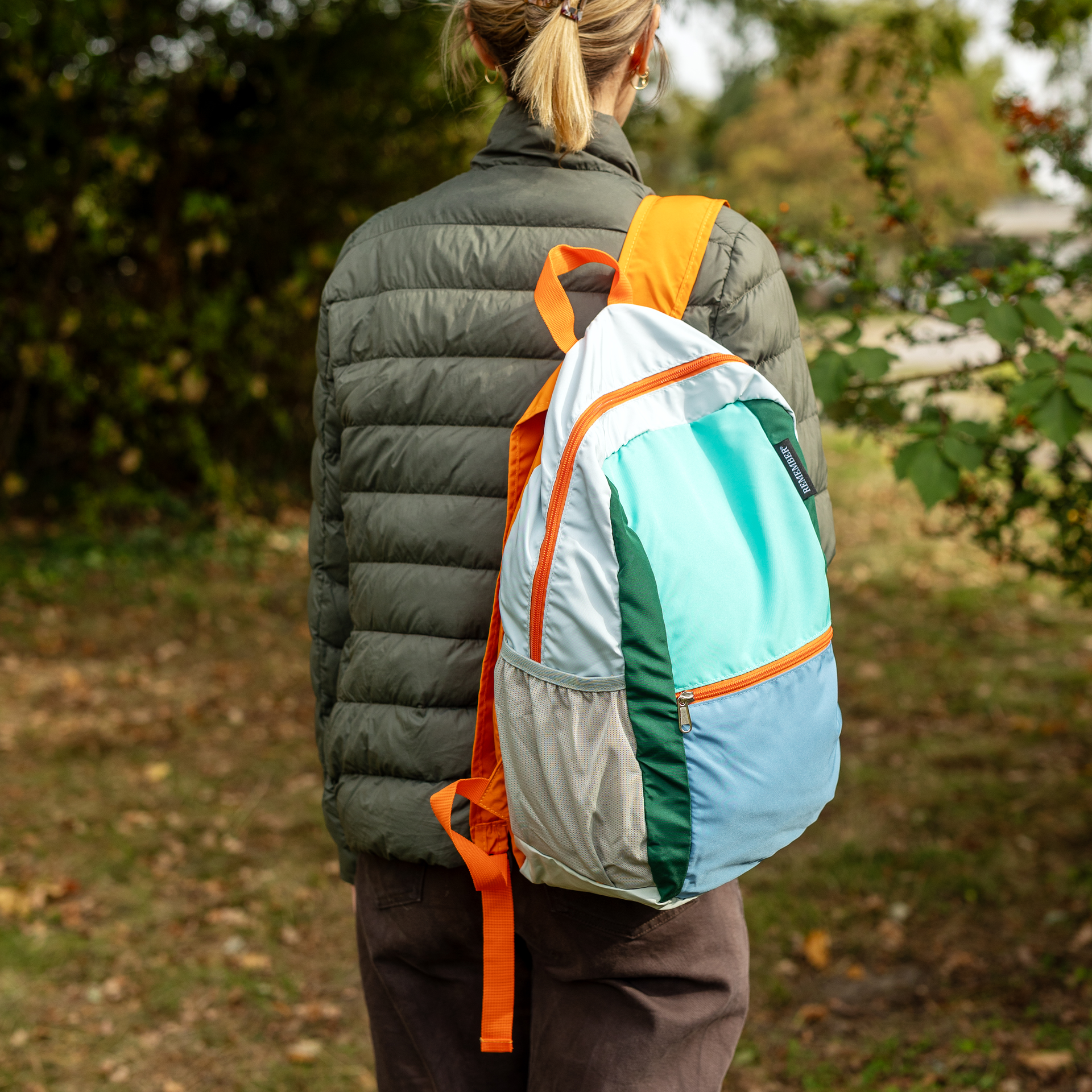 Zaino pieghevole nei colori verde menta, azzurro e verde scuro con dettagli grigio chiaro e arancione. Tasca frontale con cerniera arancione, spallacci e maniglia di trasporto arancioni, nonché tasche laterali in rete. Etichetta REMEMBER® sul lato.