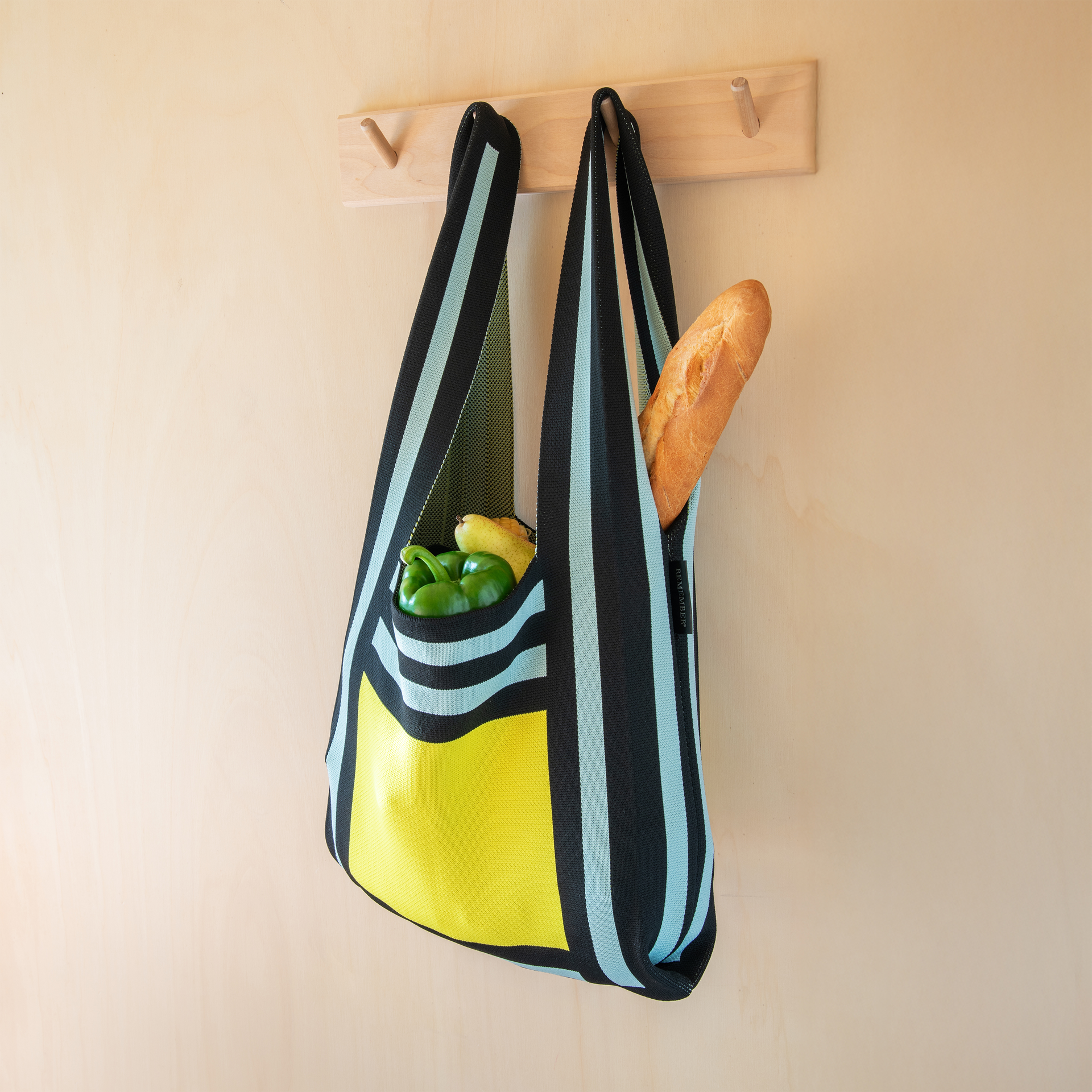 Eine gestreifte Stricktasche mit Lebensmitteln hängt an einer Garderobe. In der Tasche sind grüne Paprika, eine Birne und ein Baguette.