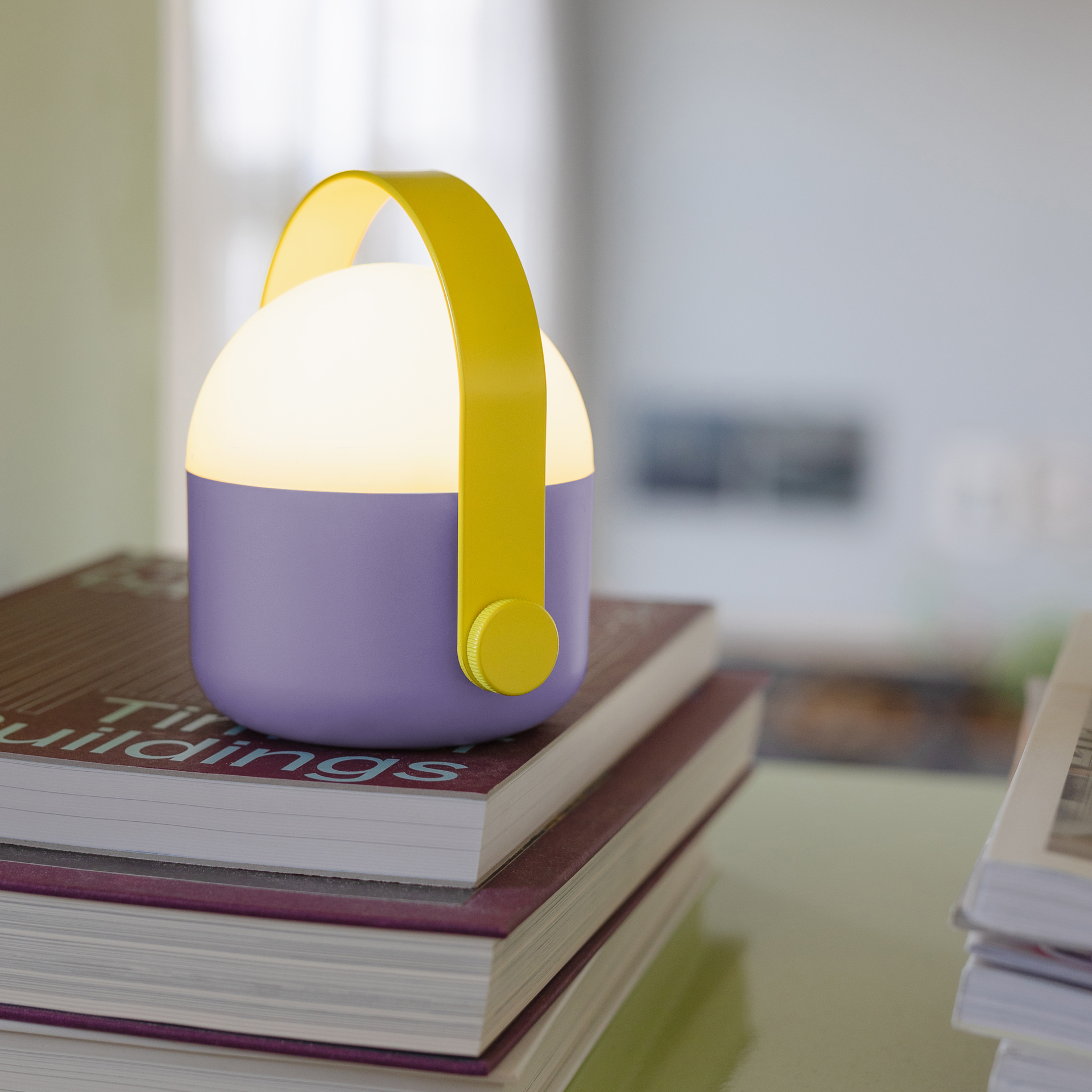 Lilafarbene Leuchte "Ole" mit gelbem Henkel, platziert auf einem Stapel Büchern. Die Leuchte strahlt warmes Licht aus.
