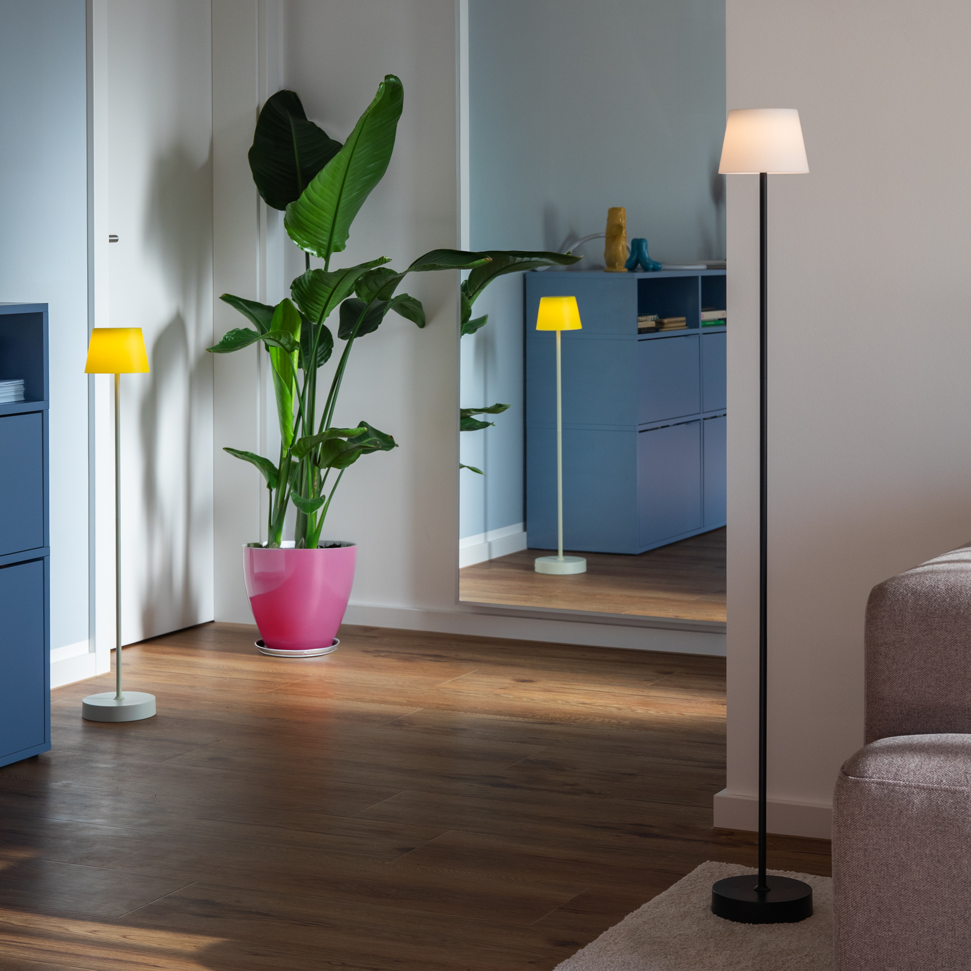 Ein Wohnraum mit braunem Holzboden, einer Stehlampe mit gelbem Schirm und einer mit weißem Schirm, einem Schrank, einer Zimmerpflanze in einem pinkfarbenen Topf und einem Spiegel, der den Raum verdoppelt.