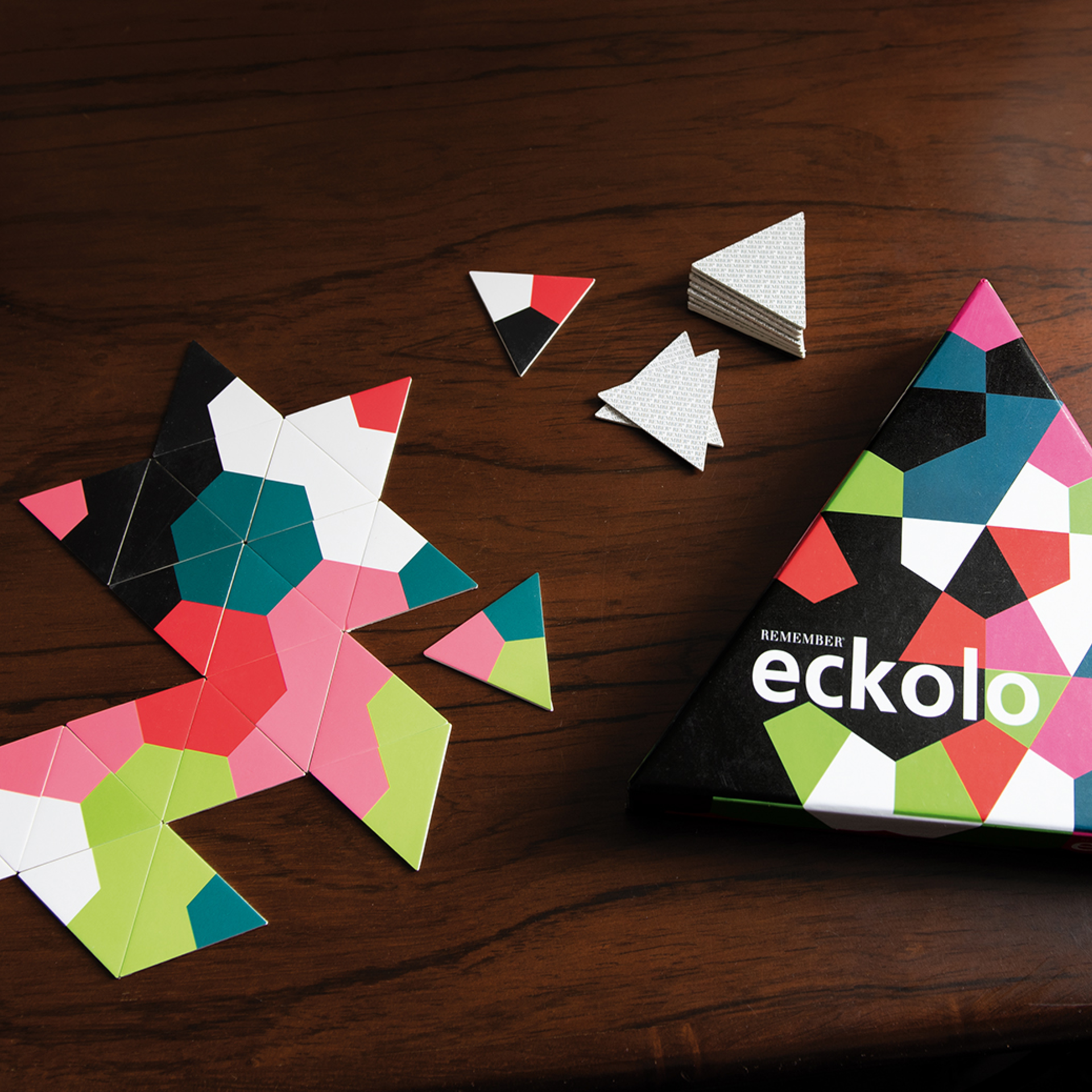 Draufsicht: Eckolo Spiel mit Dreieck-Karten, teils als Stern gelegt. Spielschachtel rechts. Kartenstapel & lose Karten. Farben: Grün, Pink, Rot, Schwarz, Weiß, Blaugrün.